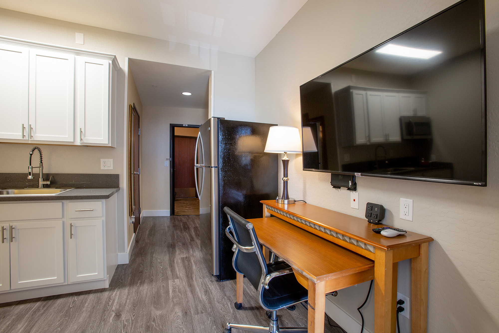 A well-lit kitchen area featuring a sink, countertop, and a black refrigerator, alongside a modern workspace with a wooden desk and chair. A large flat-screen TV is mounted on the wall, creating a cozy atmosphere. The flooring is a stylish laminate, enhancing the room's contemporary look.