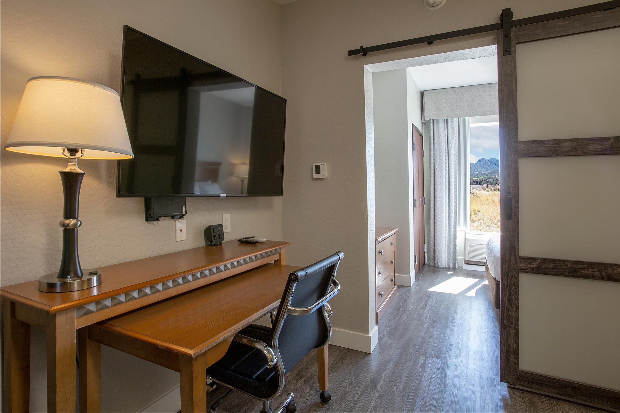 A well-lit hotel room featuring a wooden desk with a chair, a wall-mounted television, and a sliding door leading to a bright, welcoming bedroom. Natural light streams in from the window, showcasing a view of mountains outside. The interior has a modern, clean aesthetic with neutral colors.
