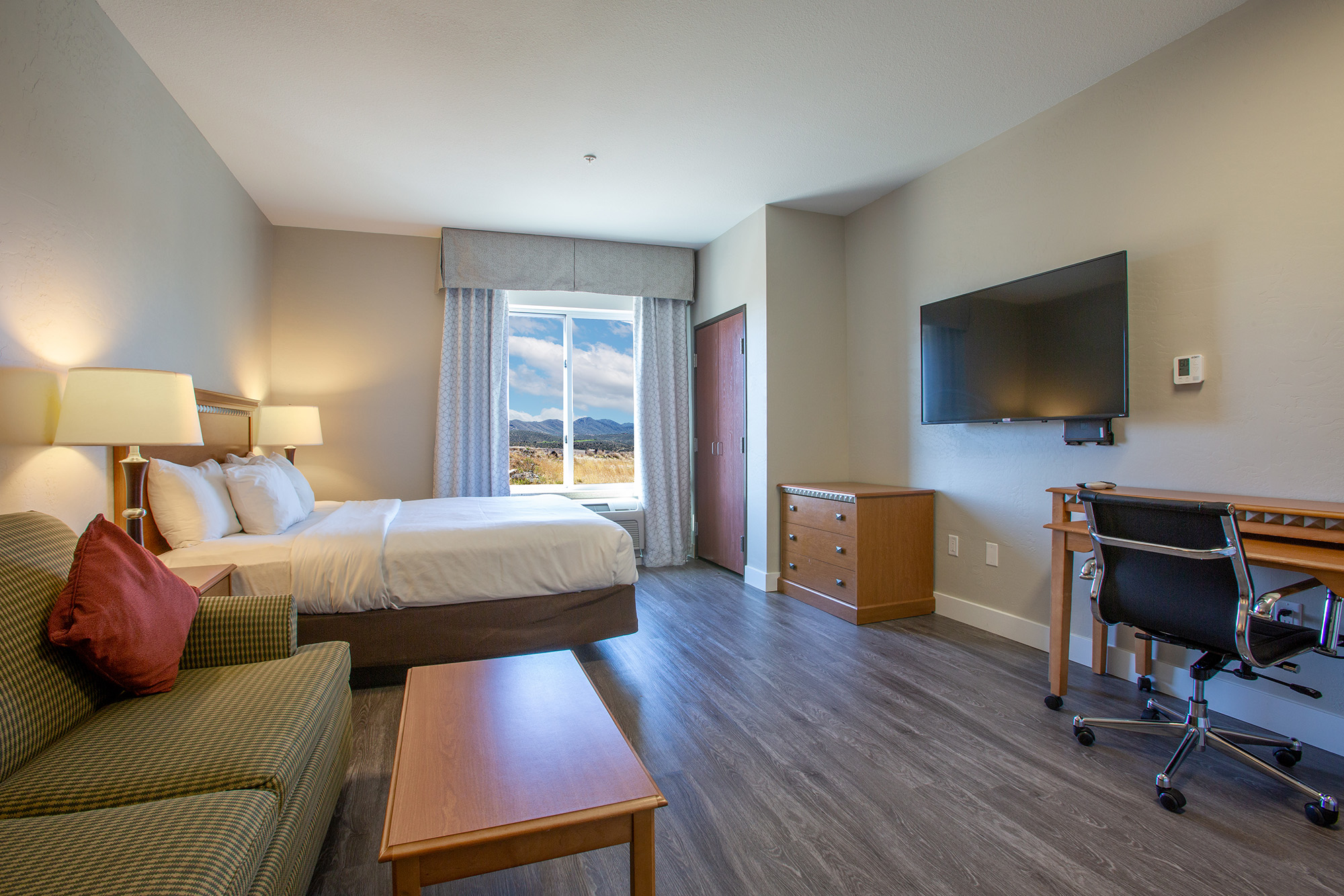 A well-lit hotel room featuring a queen bed with white linens, a comfortable green striped couch, a wooden coffee table, a desk with a chair, and a large TV mounted on the wall. A window provides a view of distant mountains and allows natural light into the space. The decor is modern and inviting.