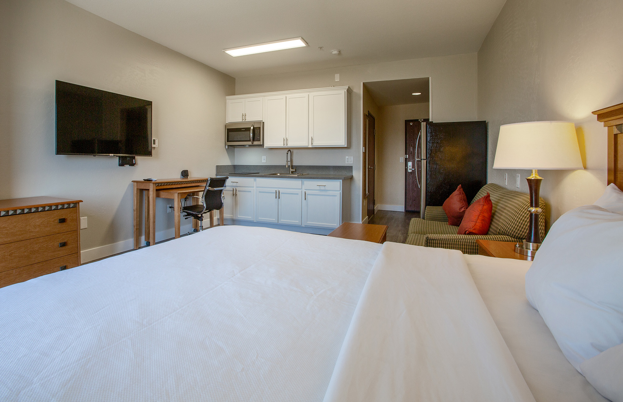 A well-furnished hotel room featuring a queen-sized bed, a kitchenette with a microwave and countertop, a flat-screen TV mounted on the wall, a small dining area with a chair, and a cozy seating space with a green couch and decorative pillows. The decor is modern and inviting.