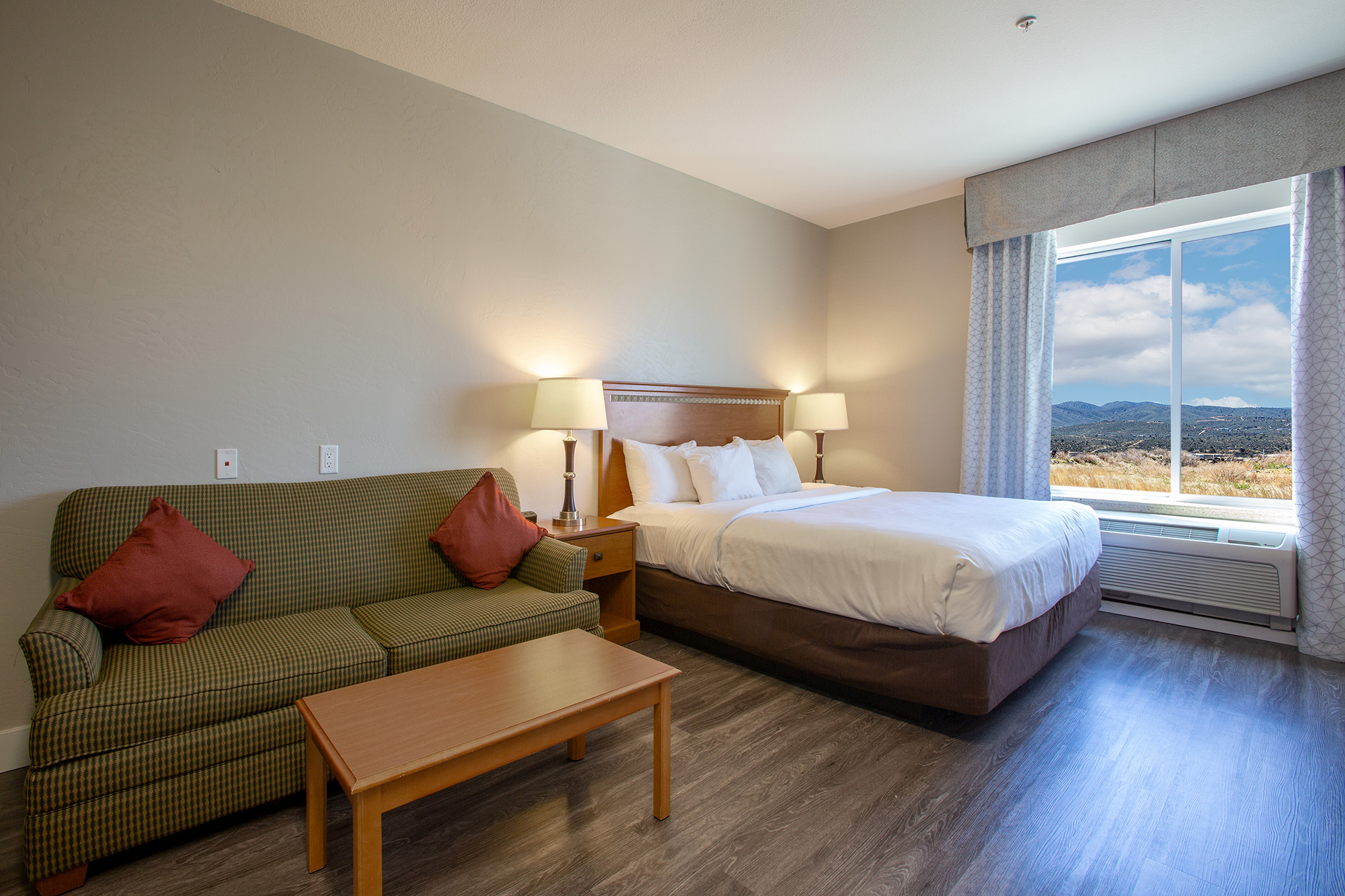A hotel room featuring a queen bed with white bedding, a green sofa with burgundy cushions, a wooden coffee table, and a window showing a scenic view of mountains and clouds. The room has soft lighting and a modern design with gray walls and a laminate floor.
