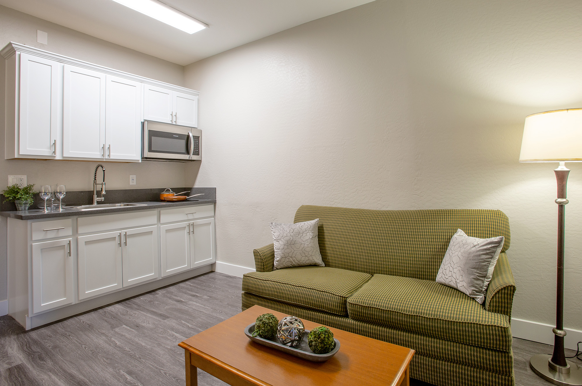 A cozy living area featuring a green checkered sofa with decorative pillows, a wooden coffee table with decorative items, a kitchenette with white cabinets, a microwave, and a light source from a floor lamp. The walls are painted a soft color, creating a warm and inviting atmosphere.