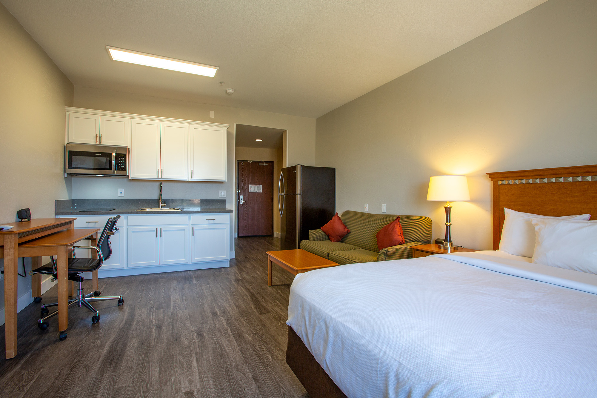 A modern hotel room featuring a bed, a kitchenette with a microwave, a refrigerator, and a dining table. The room includes a small seating area with a sofa and a lamp, along with a desk and a door leading to the hallway. Soft lighting creates a welcoming atmosphere.