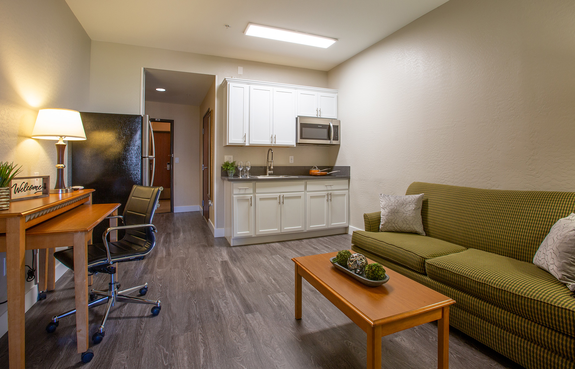 A modern living space featuring a green-striped sofa, a wooden coffee table with decorative plants, a small desk with a chair, and a kitchenette with white cabinets. The room has neutral walls, a laminate floor, and bright overhead lighting, creating a welcoming atmosphere.