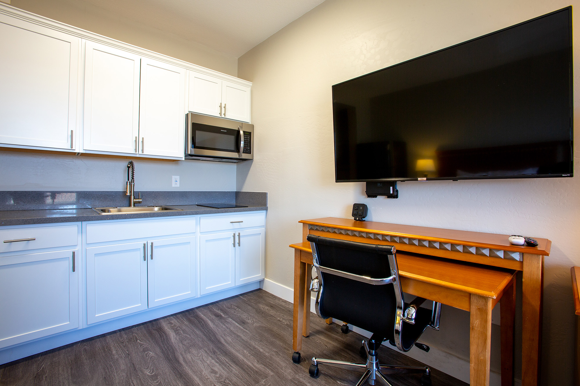 A modern kitchen area featuring white cabinets, a microwave, and a sink. To the right, there is a large flat-screen TV mounted on the wall. Below the TV is a wooden desk with a chair, providing a workspace. The floor is a dark laminate, adding to the contemporary look of the space.