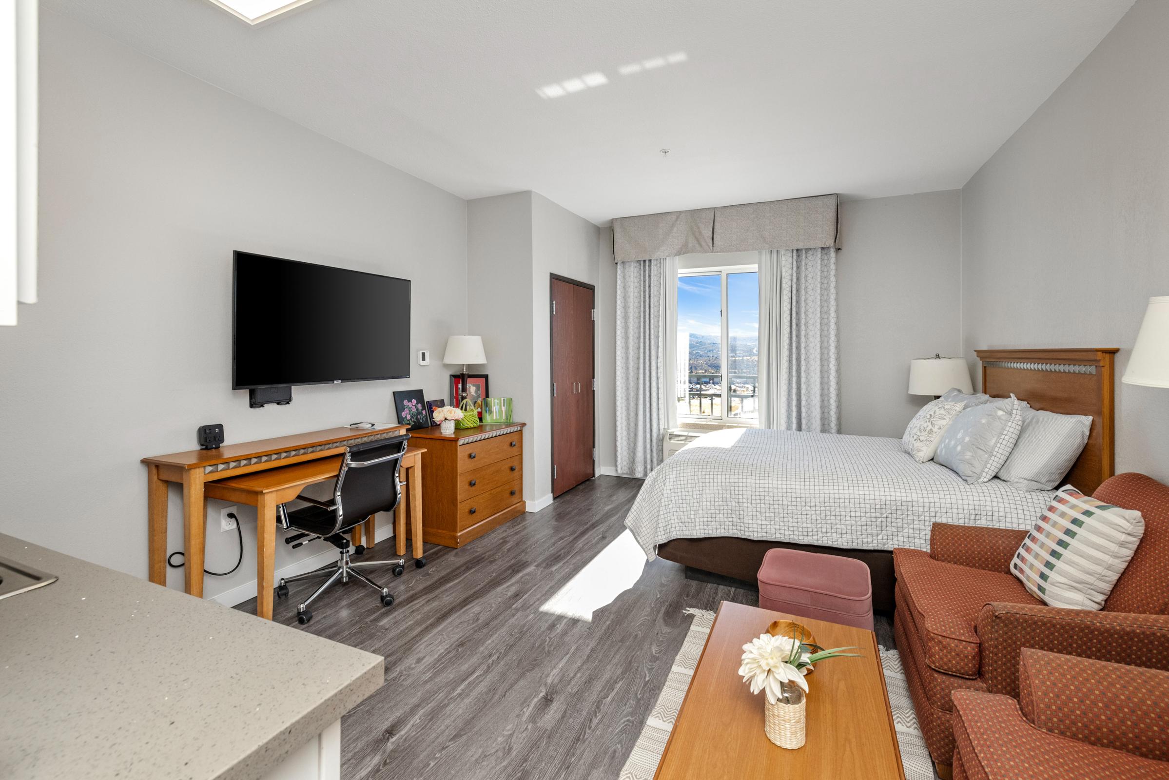 A modern hotel room featuring a queen bed with decorative pillows, a small dining area, a flat-screen TV mounted on the wall, a desk with a chair, and a window offering views. The room has light-colored walls and wooden furniture, creating a cozy and inviting atmosphere.