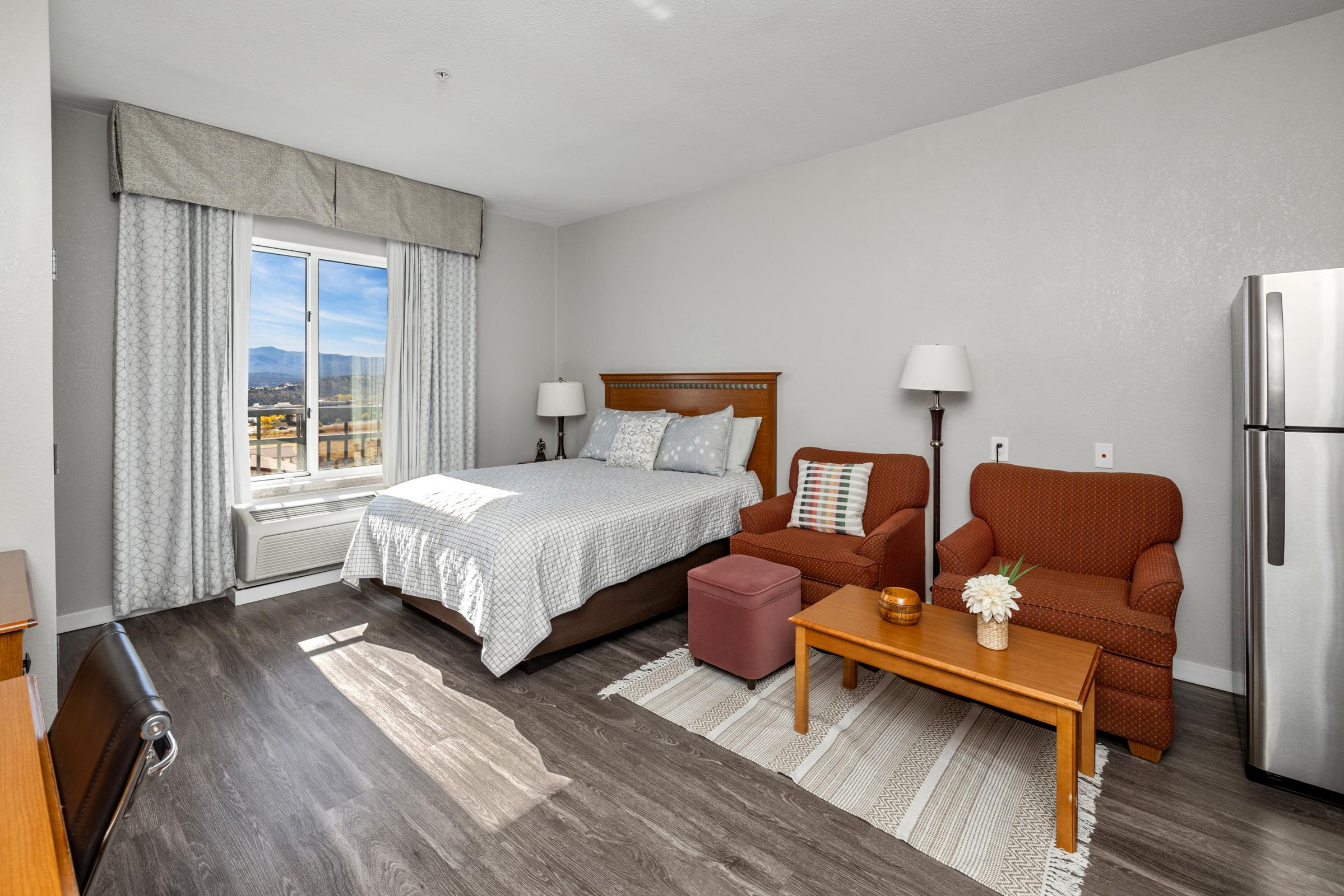 A well-lit hotel room featuring a queen-sized bed with decorative pillows, two armchairs, a small coffee table, and a mini-fridge. Large windows reveal a scenic view of mountains. Soft gray walls and wooden flooring create a modern and inviting atmosphere.