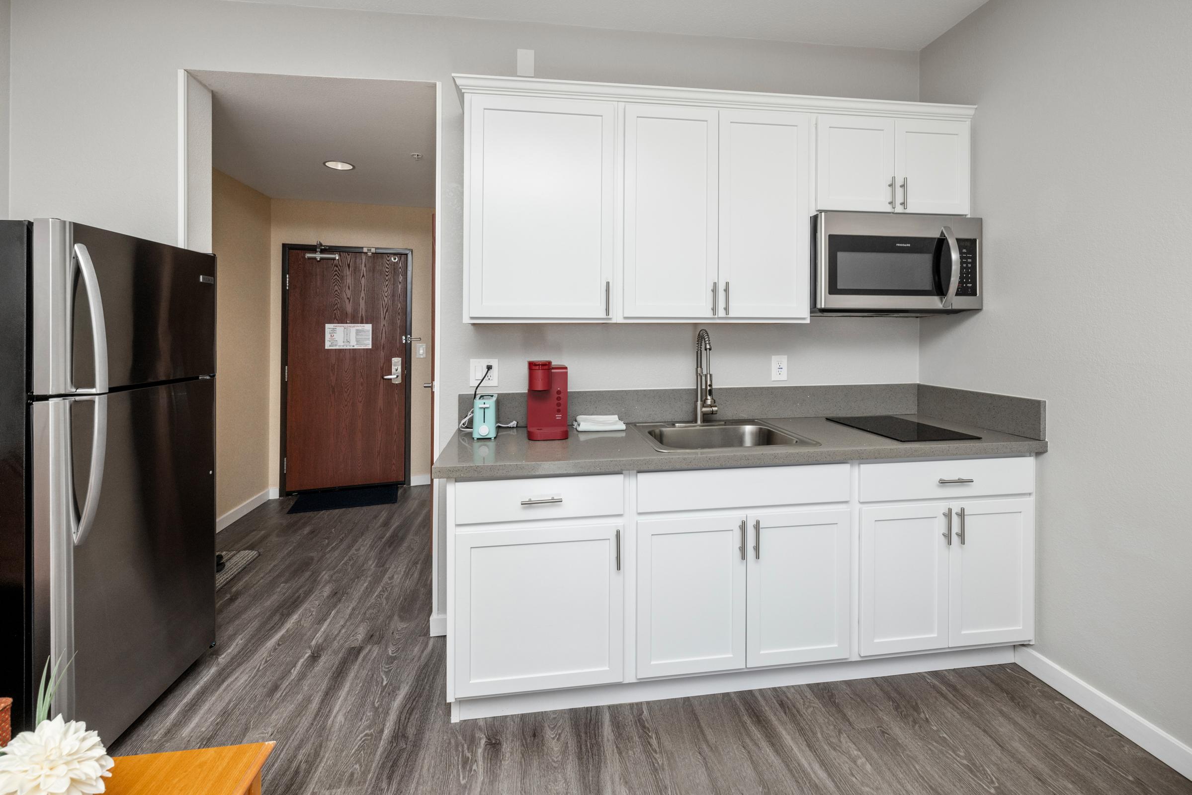 Modern kitchen featuring white cabinets, a stainless steel refrigerator, a microwave, and a sink. A coffee maker is on the countertop. The floor is hardwood, and there is a glimpse of a doorway leading to another room. The overall color scheme is light and contemporary.