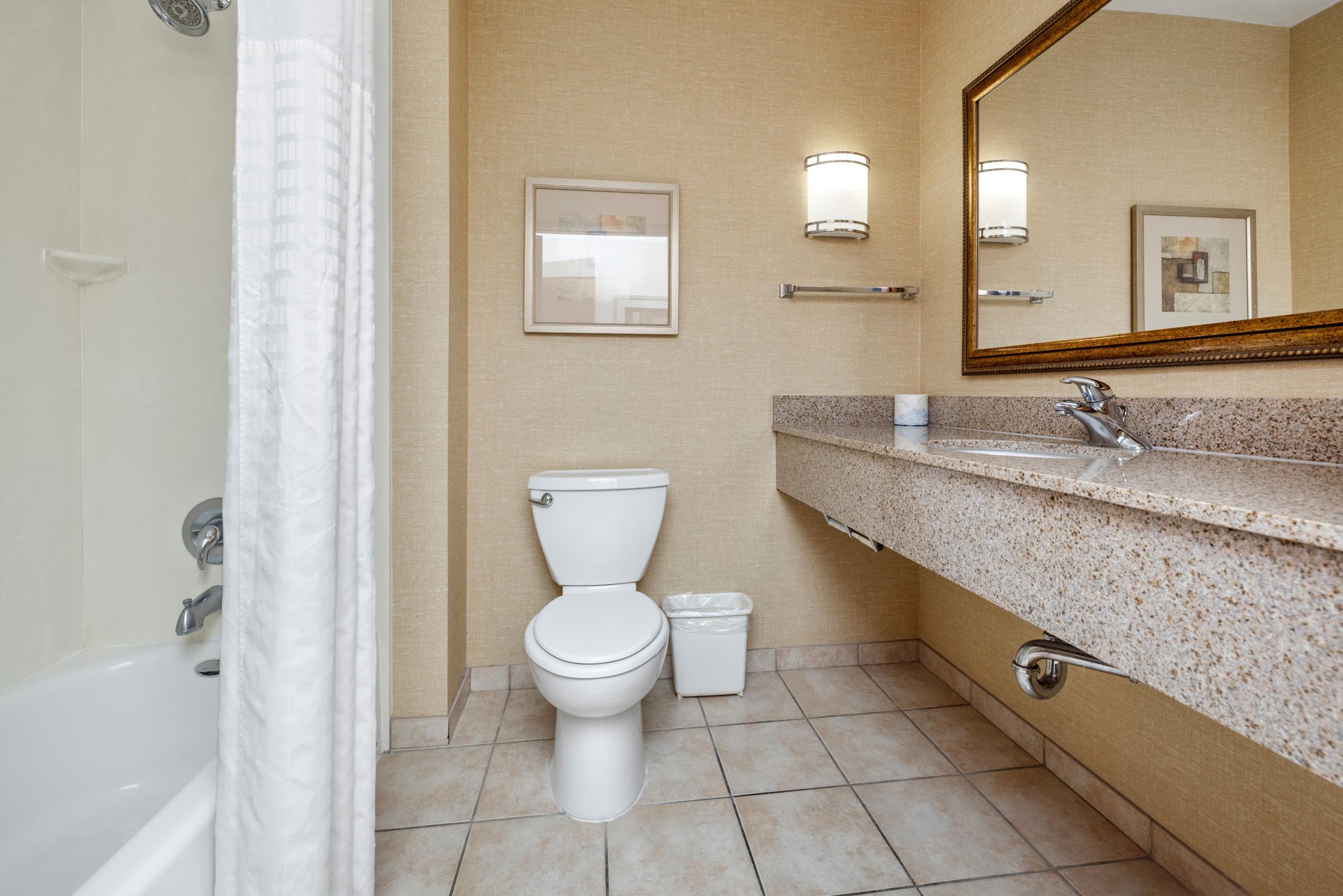 Clean and modern bathroom featuring a white toilet, a bathtub with a shower curtain, a large granite countertop with a sink, and a wall-mounted mirror. The walls are covered in a light beige texture, and there are two wall sconces providing ambient lighting. A small trash bin is placed beside the sink.