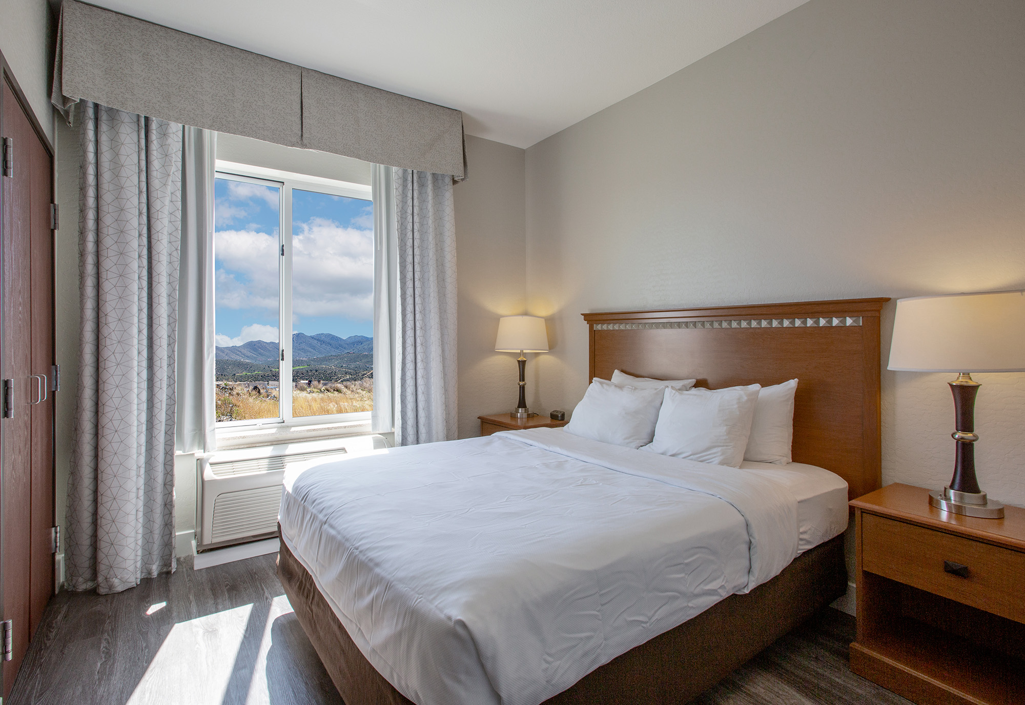 A cozy hotel room featuring a queen bed, bedside tables with lamps, and a large window offering a view of mountains. The walls are painted in neutral tones, and the room is well-lit with natural light, creating a welcoming atmosphere.