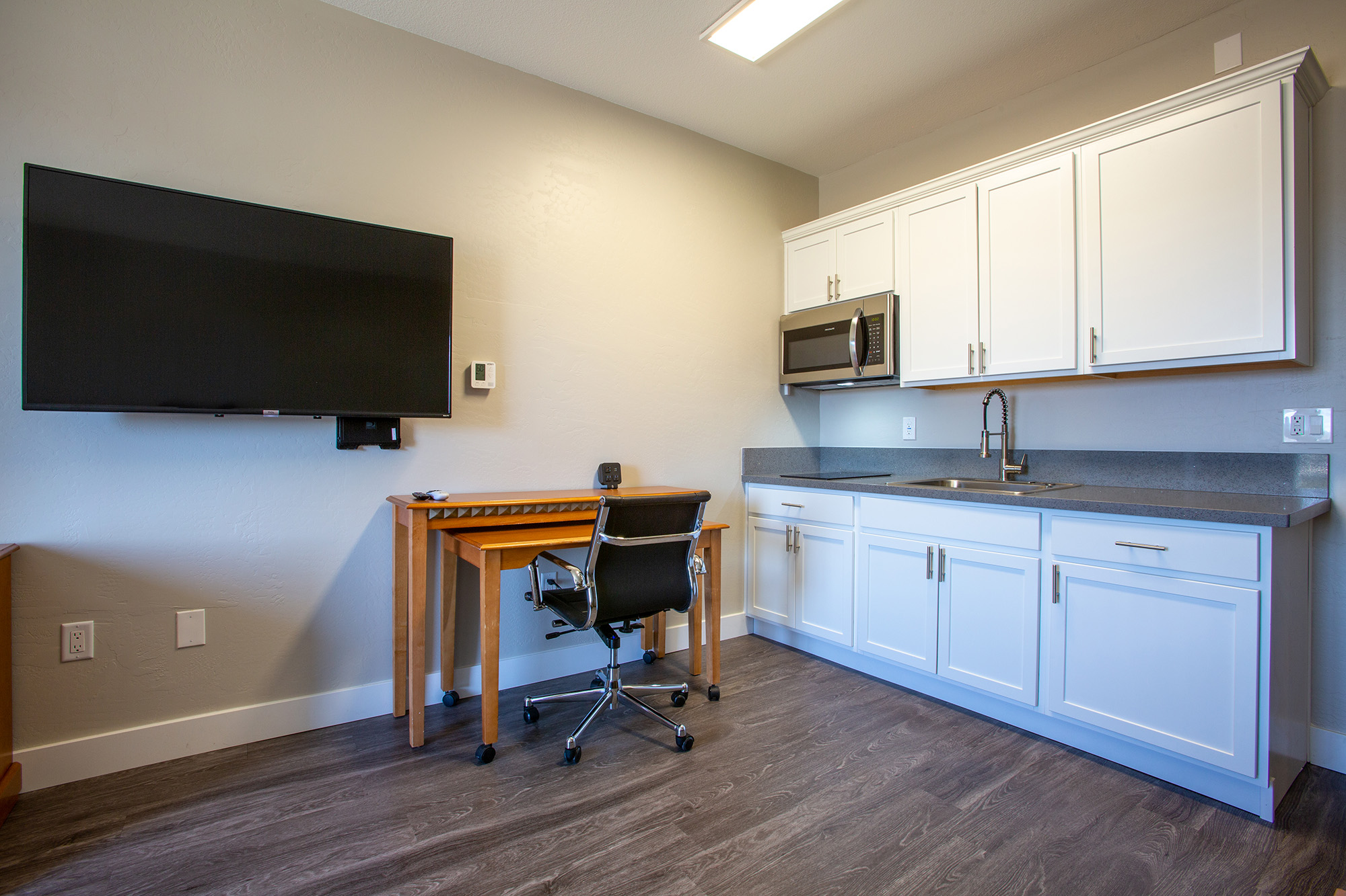 A modern studio apartment featuring a small kitchenette with white cabinets, a microwave, and a sink. A wooden desk with a black rolling chair is positioned beside the kitchenette. A large flat-screen TV is mounted on the wall opposite the desk, and the floor has a wood-like finish.