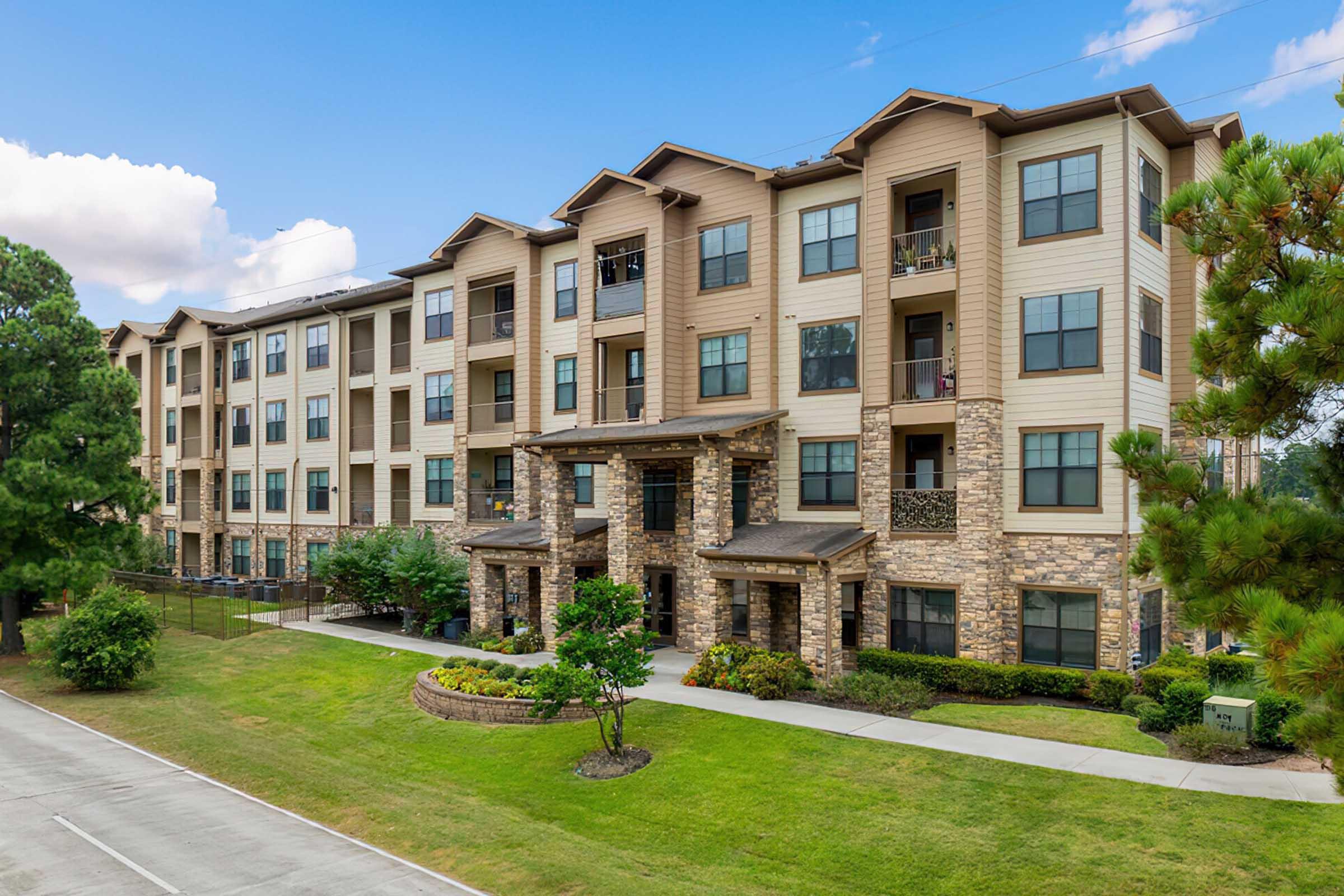 A modern multi-story apartment building featuring a combination of stone and siding exteriors. The structure is landscaped with trees and shrubs, and there is a paved pathway in front. The sky is partly cloudy, creating a vibrant atmosphere.