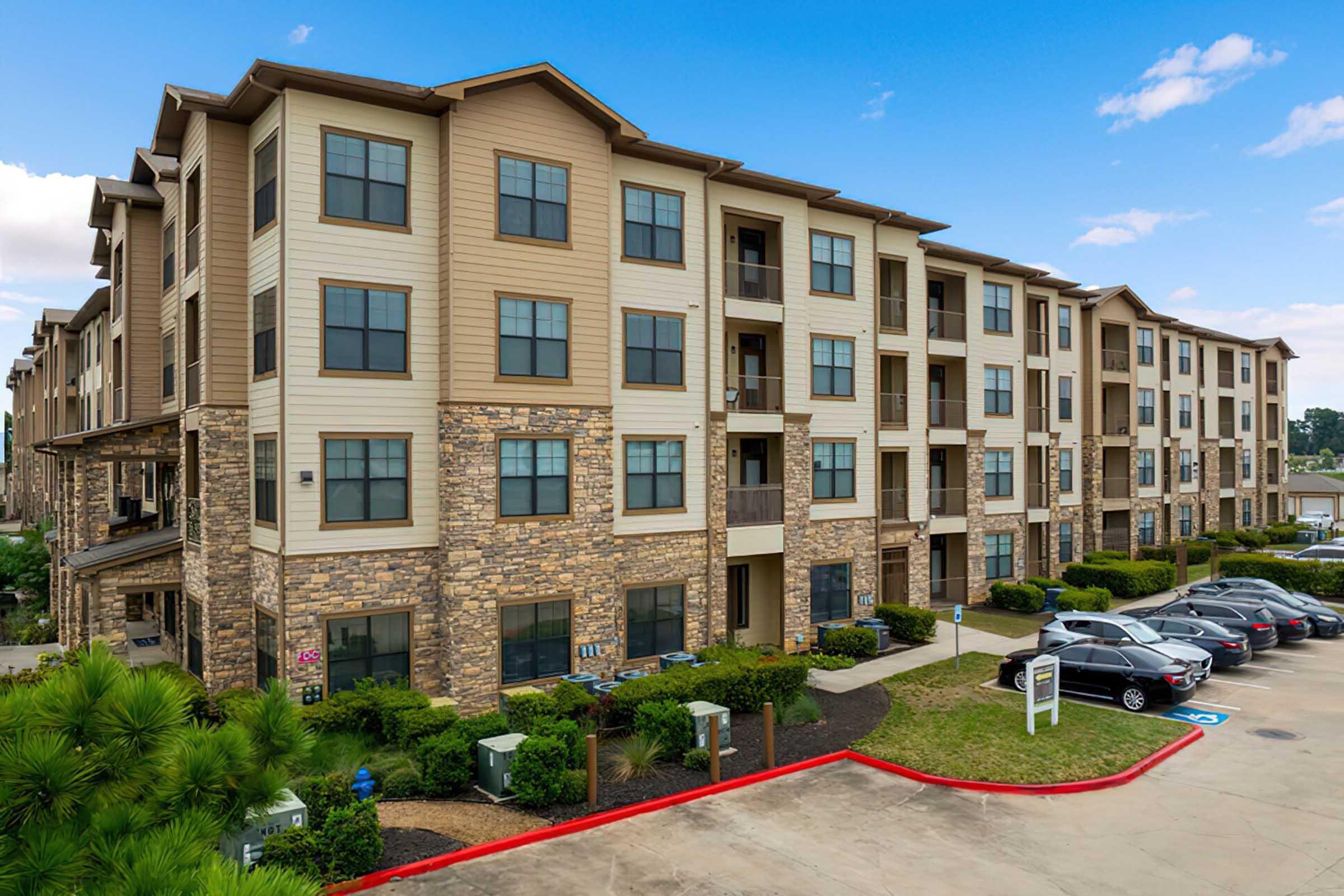 A multi-story residential building featuring a blend of stone and wood siding. The architecture includes numerous windows and balconies. In the foreground, there is a parking lot with several cars and landscaped greenery. The sky is clear with a few clouds, creating a bright atmosphere.