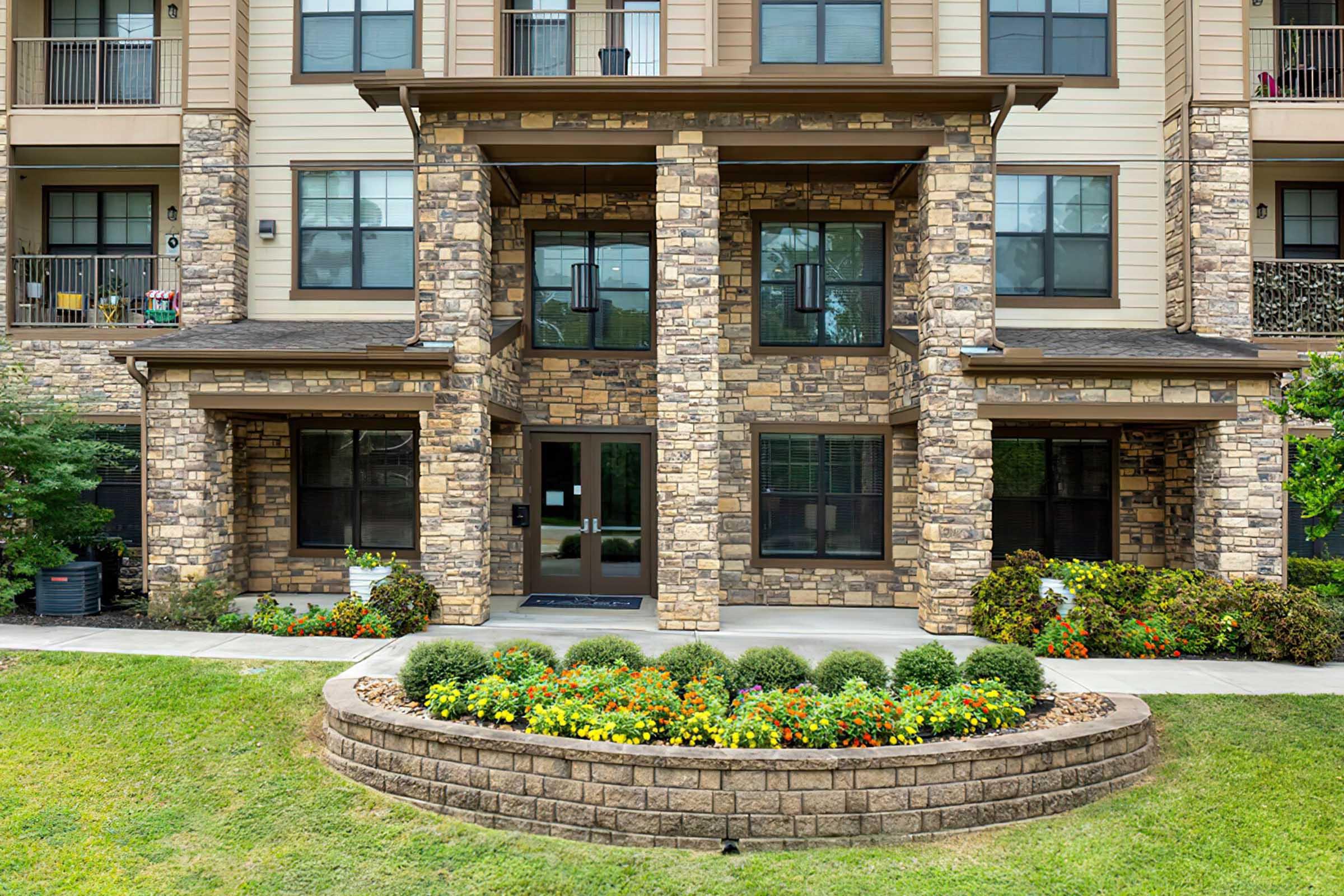 A modern apartment building featuring stone and beige siding. The entrance is framed by two large columns, with glass doors and a welcoming stone walkway. Colorful flower beds and well-maintained greenery enhance the front yard, adding to the inviting atmosphere.