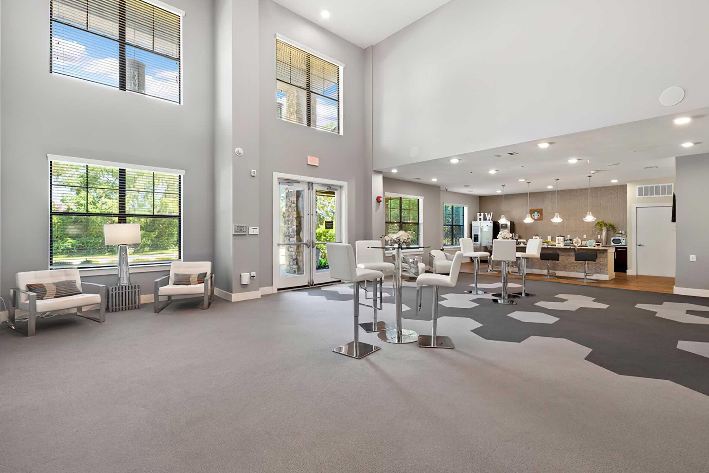 Bright and spacious lounge area featuring modern furniture, large windows allowing natural light, and a stylish kitchen space in the background. The floor is covered in a patterned carpet, and the decor is contemporary and inviting, suitable for social gatherings.