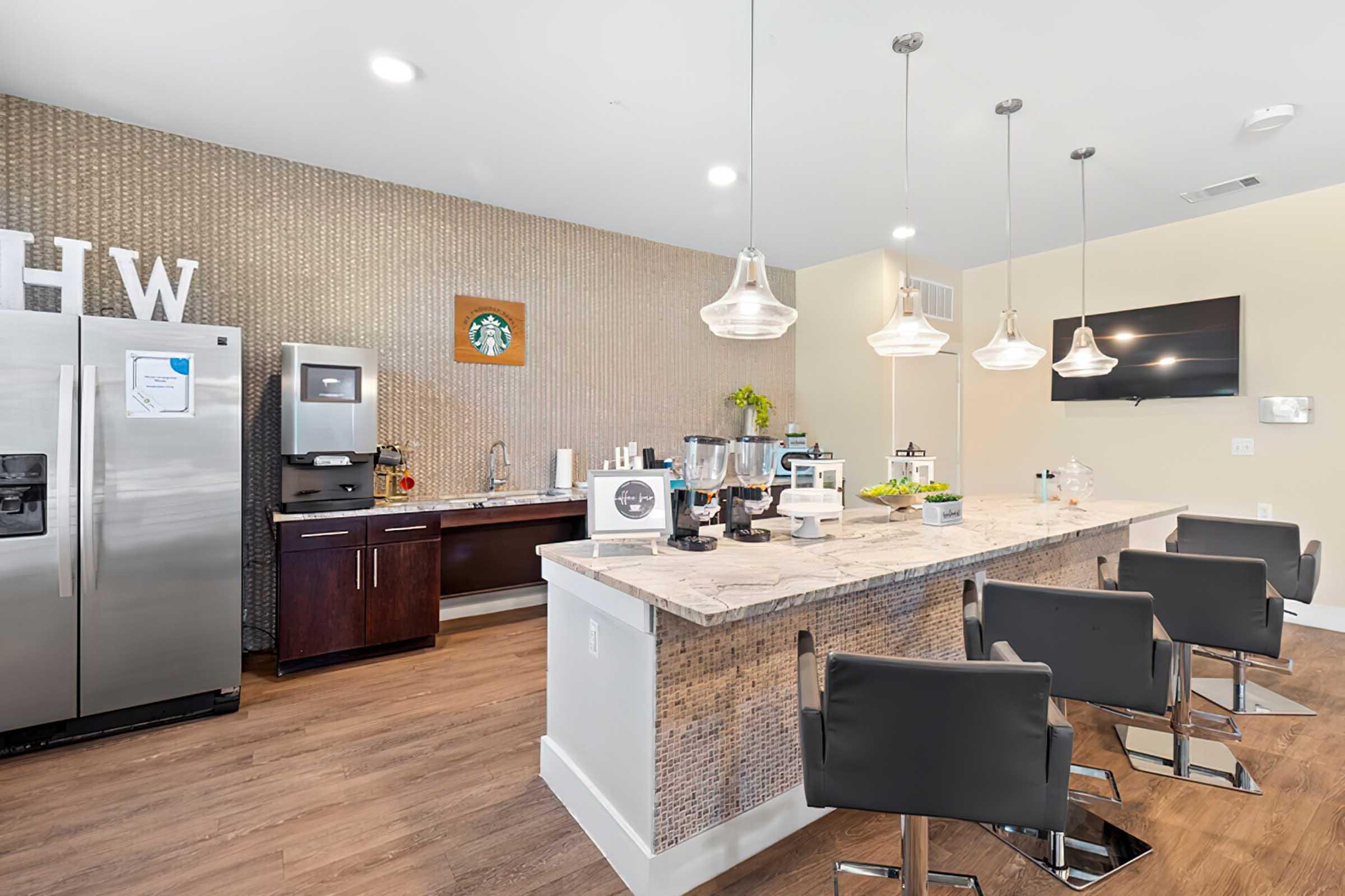 Modern kitchen with a sleek design featuring a large bar area with black chairs. A stylish backsplash complements stainless steel appliances, including a refrigerator and coffee machine. Natural light fills the space, with decorative elements like plants adding a fresh touch. A flat-screen TV is mounted on the wall.
