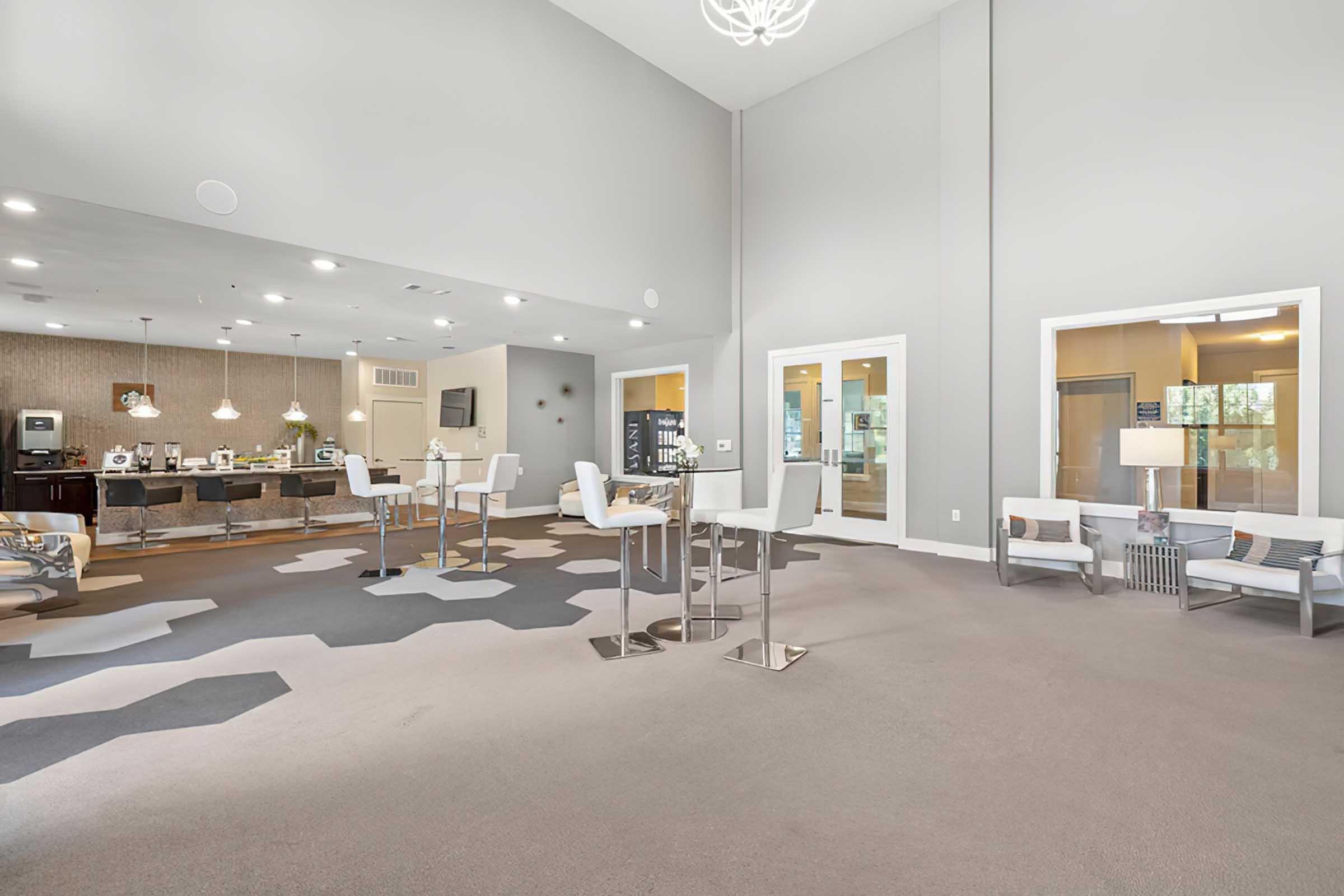 A spacious, modern interior with high ceilings and neutral-colored walls. The area features contemporary furniture, including white high chairs and tables, and gray and beige patterned carpet. In the background, a well-lit kitchenette area is visible, enhancing the inviting atmosphere.