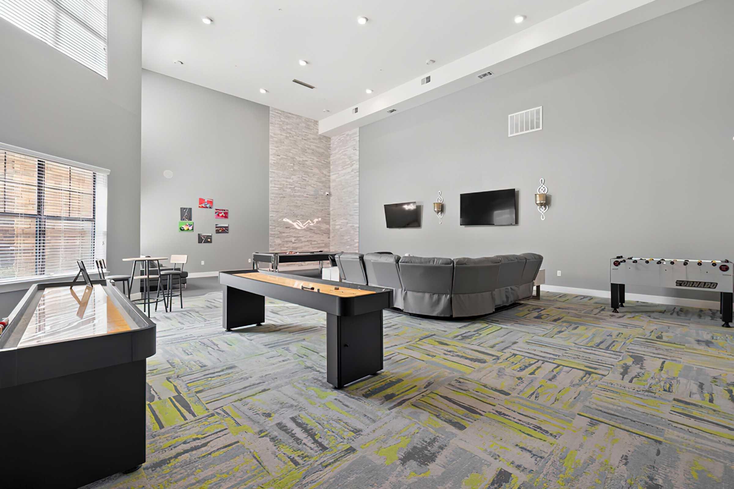 A spacious lounge area featuring modern furniture, including a large gray sectional sofa, two wall-mounted TVs, a foosball table, and a game table. The room has high ceilings, natural light from large windows, and a contemporary gray and green patterned carpet. Wall art adds a touch of color to the space.