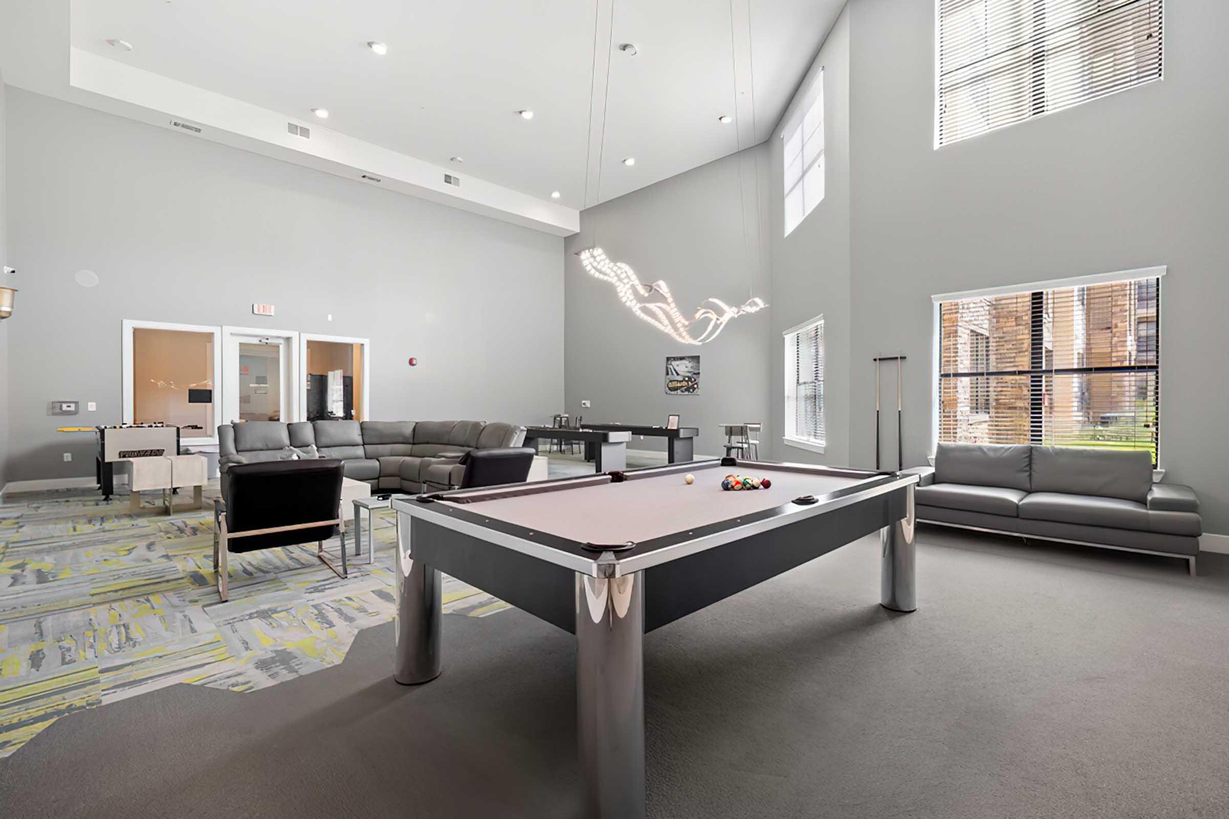 A modern recreational room featuring a billiards table in the center, surrounded by comfortable seating and large windows. The walls are painted in light gray, with stylish artwork and contemporary lighting fixtures that enhance the inviting atmosphere.