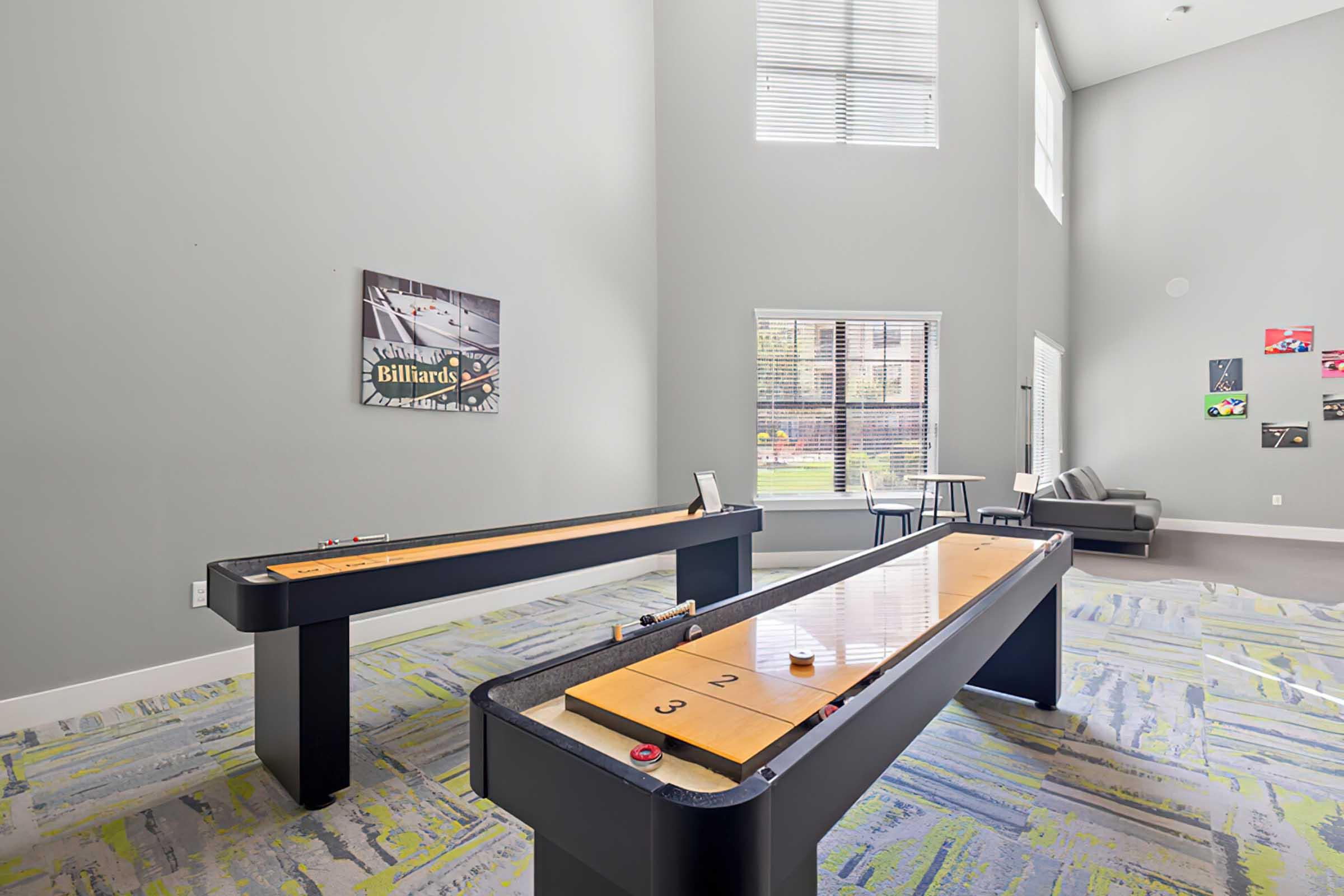 A bright game room featuring two shuffleboard tables set against a gray wall with large windows. One side has a small seating area with a sofa, while the other displays a billiards poster. The floor is covered with a colorful, patterned carpet, enhancing the inviting atmosphere.