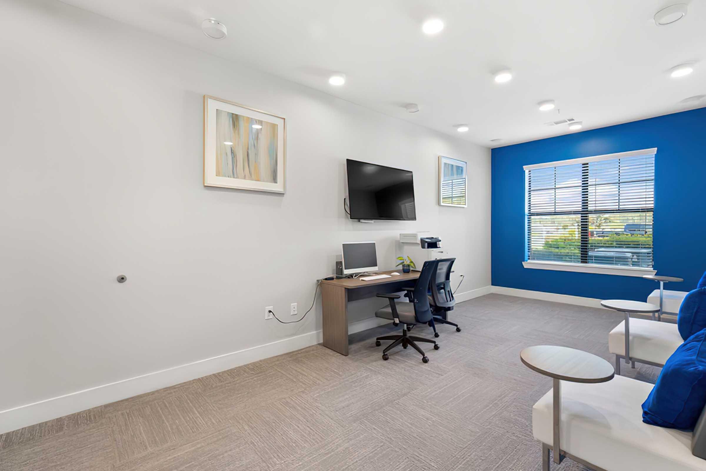 A modern office space featuring a desk with a computer, a comfortable office chair, and a wall-mounted TV. There are two small side tables and a large window with blinds, letting in natural light. The walls are painted in a calming light gray with one accent wall in deep blue. Artwork is displayed on the wall.