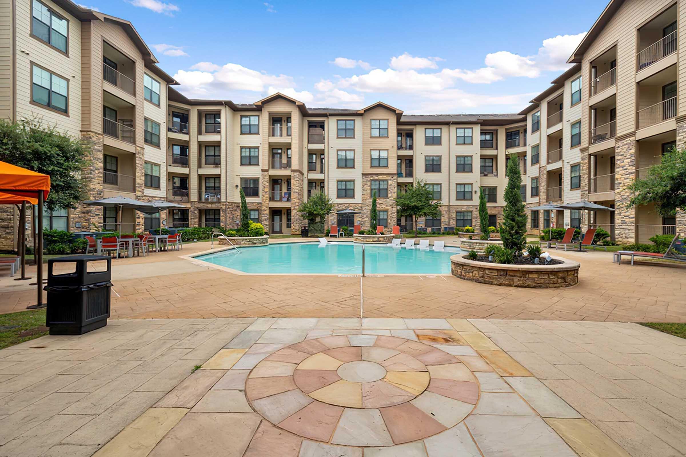 A serene apartment complex courtyard featuring a large swimming pool surrounded by lounge chairs and umbrellas. Lush greenery and well-maintained landscaping complement the modern, multi-story buildings in the background under a clear blue sky.