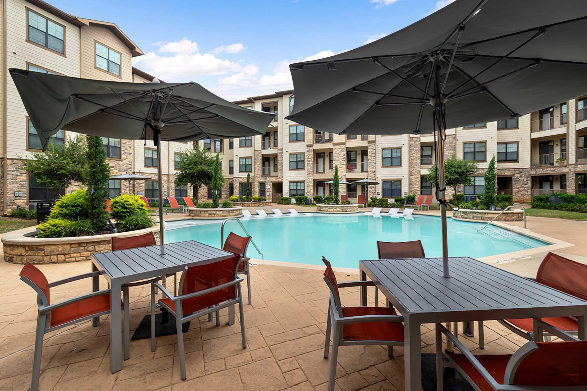 A serene pool area surrounded by apartment buildings, featuring a large swimming pool with lounge chairs and vibrant umbrellas. Tables with chairs are arranged nearby, set against a backdrop of lush greenery and clear blue skies. Ideal for relaxation and socializing.