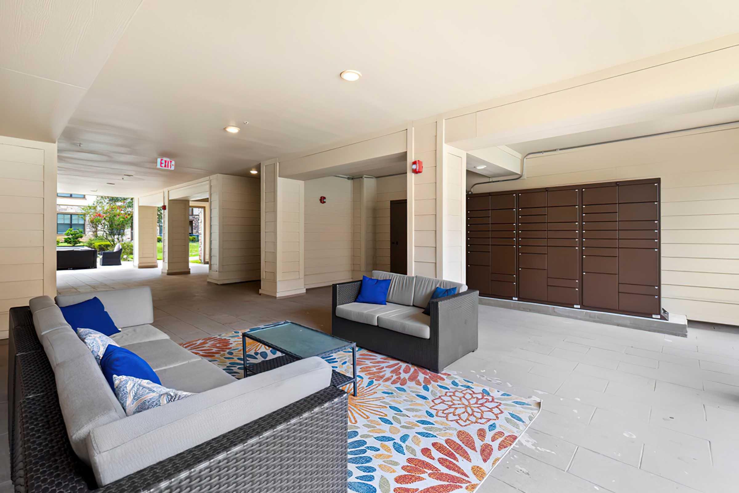 A covered outdoor area featuring a seating arrangement with two sofas adorned with blue cushions, a glass coffee table, and a colorful floral-patterned rug. In the background, there's a row of mailboxes against a light-colored wall, with green landscaping visible outside.