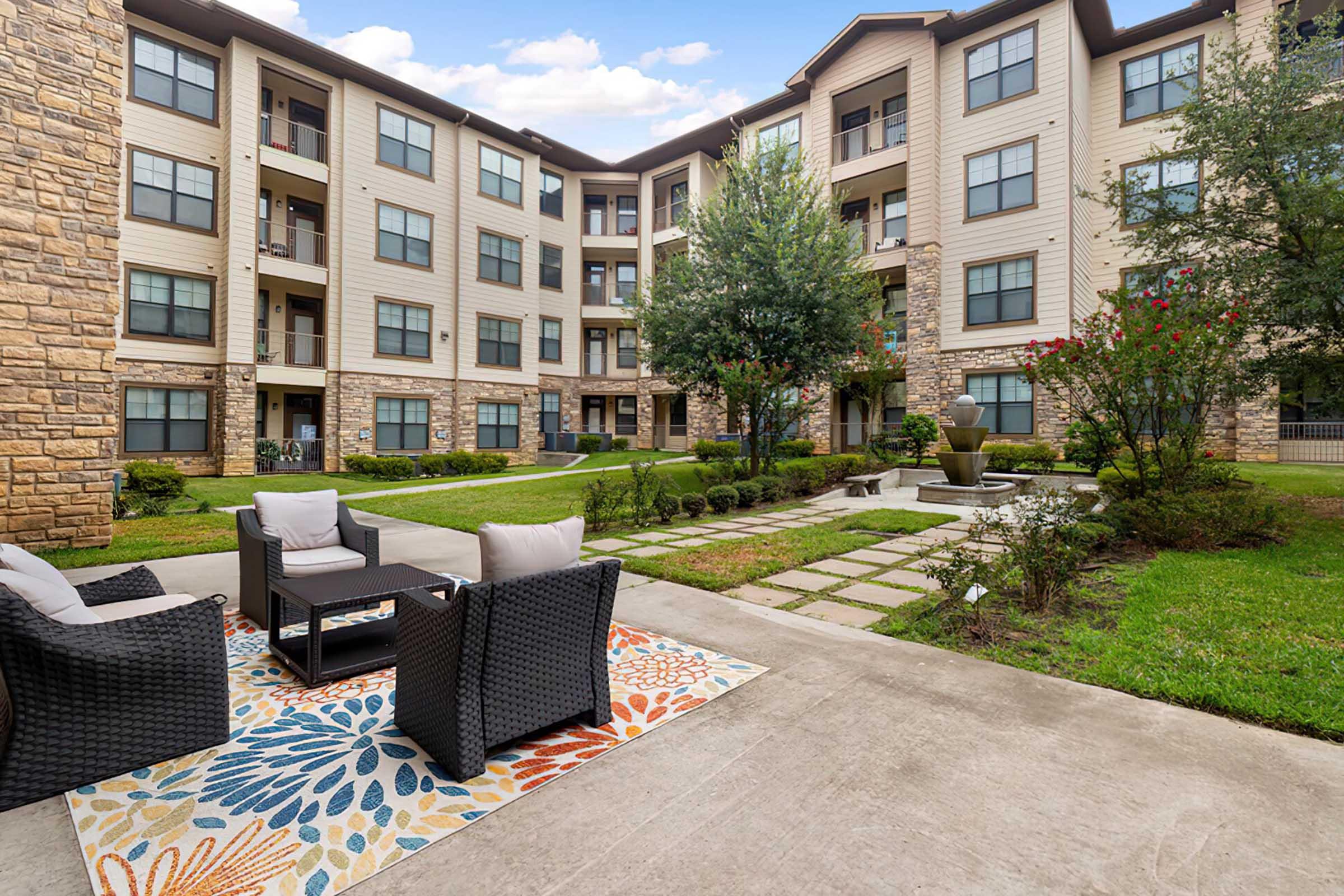 A well-maintained courtyard with modern outdoor seating and a colorful area rug, surrounded by landscaped greenery and flower beds. In the background, there are multi-story residential buildings with a mix of stone and wood exteriors. The scene is bright and inviting, ideal for relaxation.