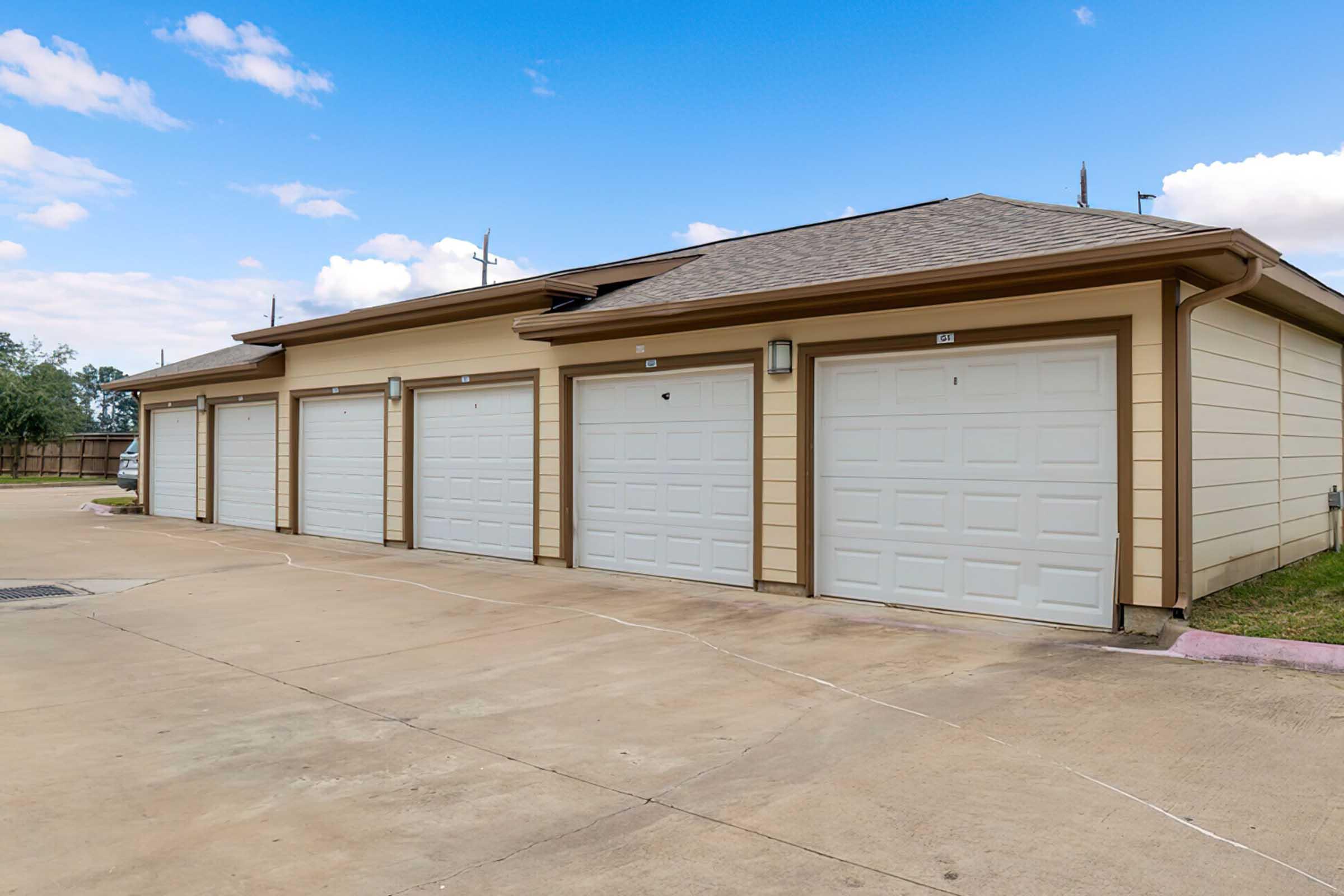 A row of five identical garage doors with white panels, situated in a paved area. The building has a light brown exterior, with a sloped roof and a clear blue sky in the background. There are no vehicles visible, and the surroundings appear tidy and well-maintained.