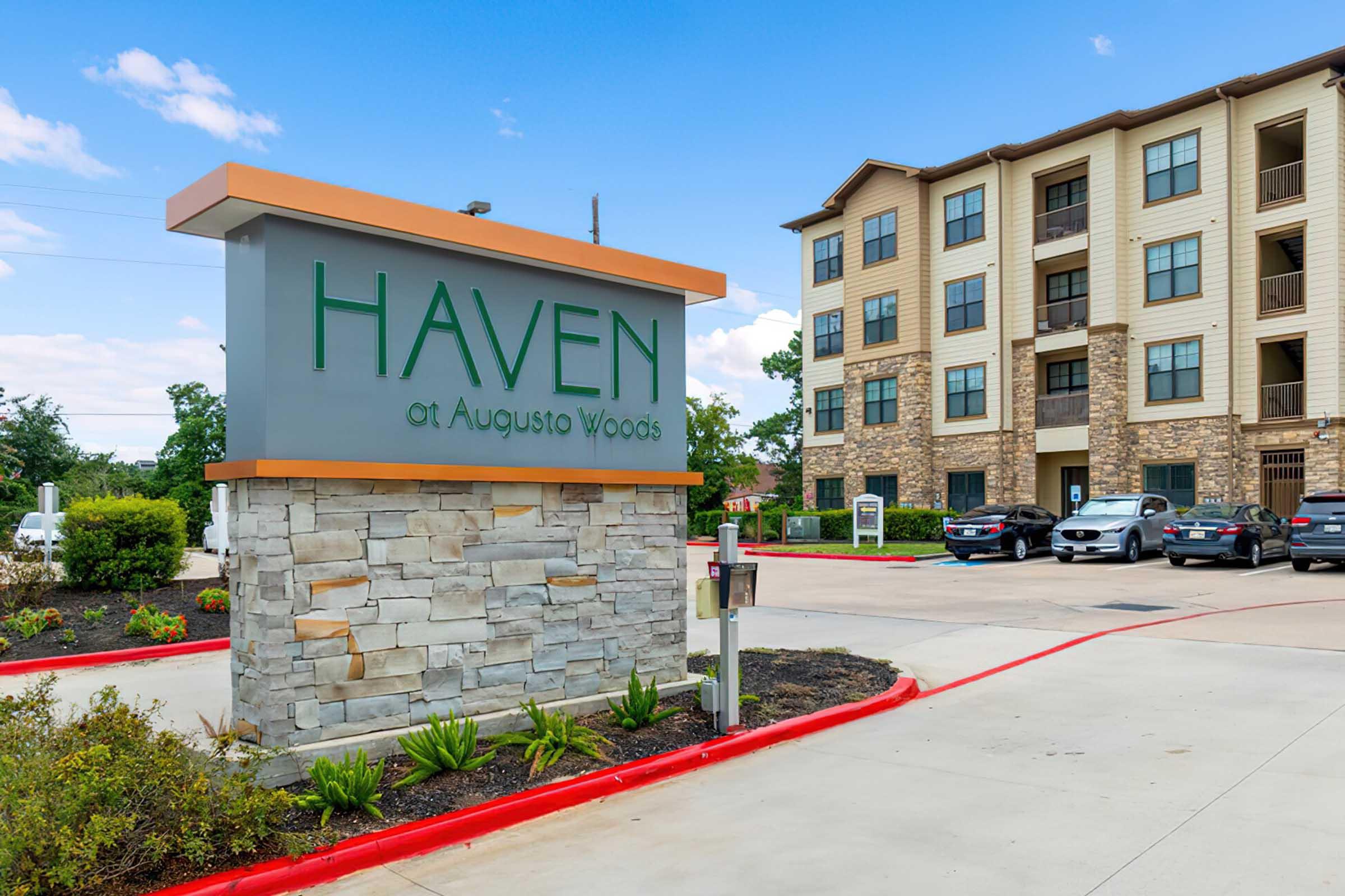 Sign for "Haven at Augusto Woods" with a modern design, featuring stonework and greenery. In the background, an apartment building is visible, showcasing a mix of materials. The entrance area has a paved driveway and parking spaces. Bright blue sky with scattered clouds.