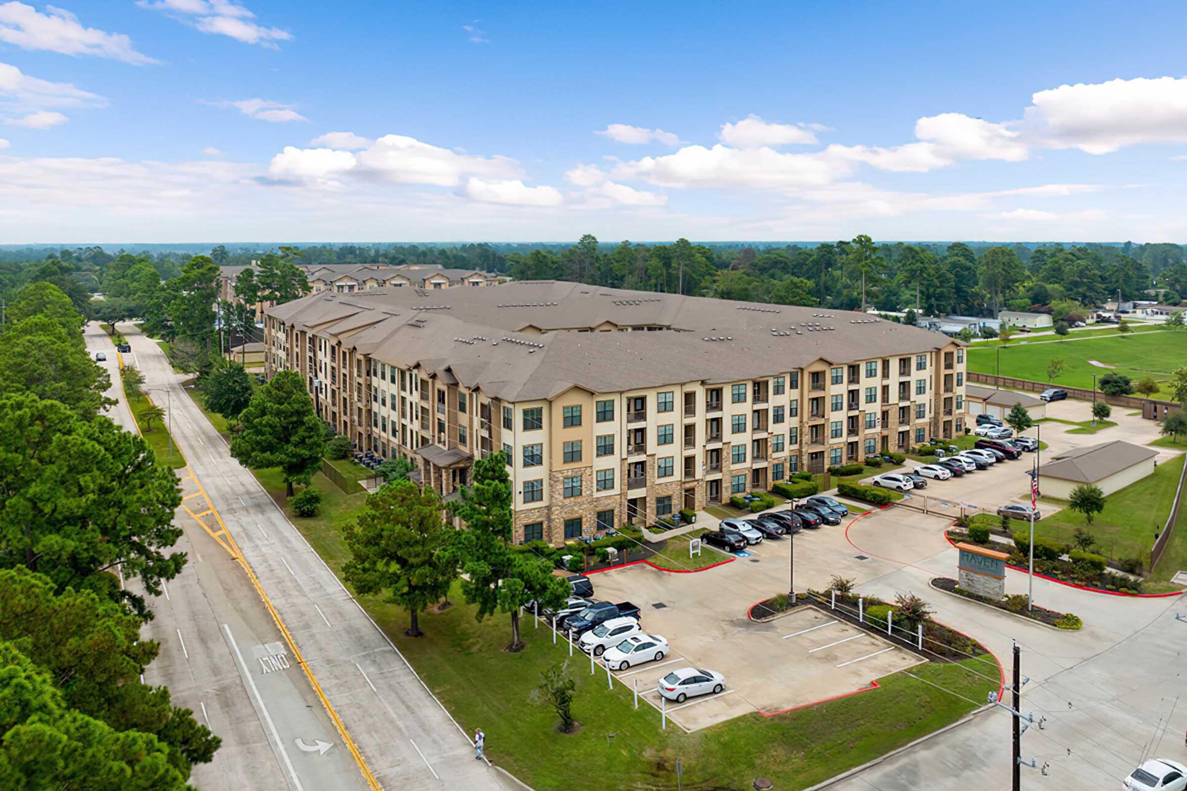 Aerial view of a multi-story residential building surrounded by trees, with a parking lot filled with cars. The building features a modern design with balconies and large windows. A wide road runs alongside it, leading to a green area in the background under a clear blue sky.