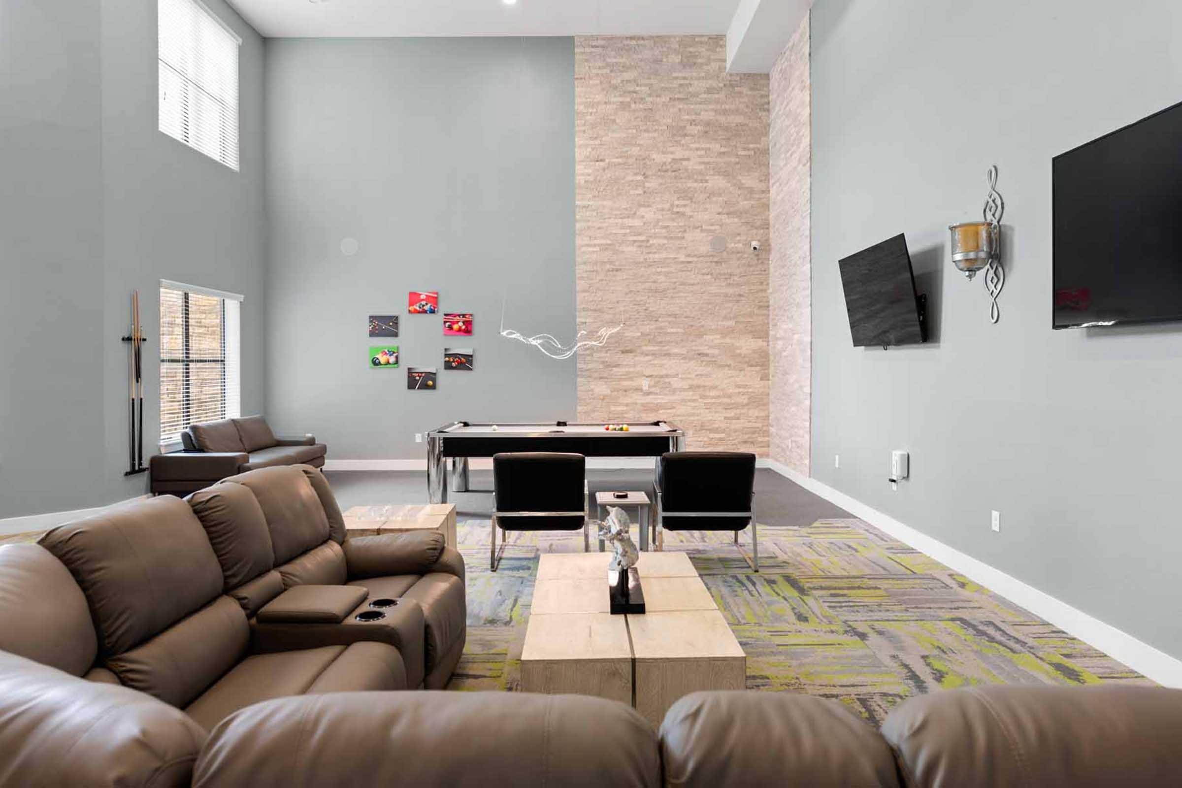 A spacious lounge area with gray walls, featuring a stone accent wall. There are two large flat-screen TVs mounted, a billiards table, and a coffee table in the center. Comfortable brown leather sofas are arranged for seating, and colorful artwork is displayed on the wall. Natural light comes through windows.