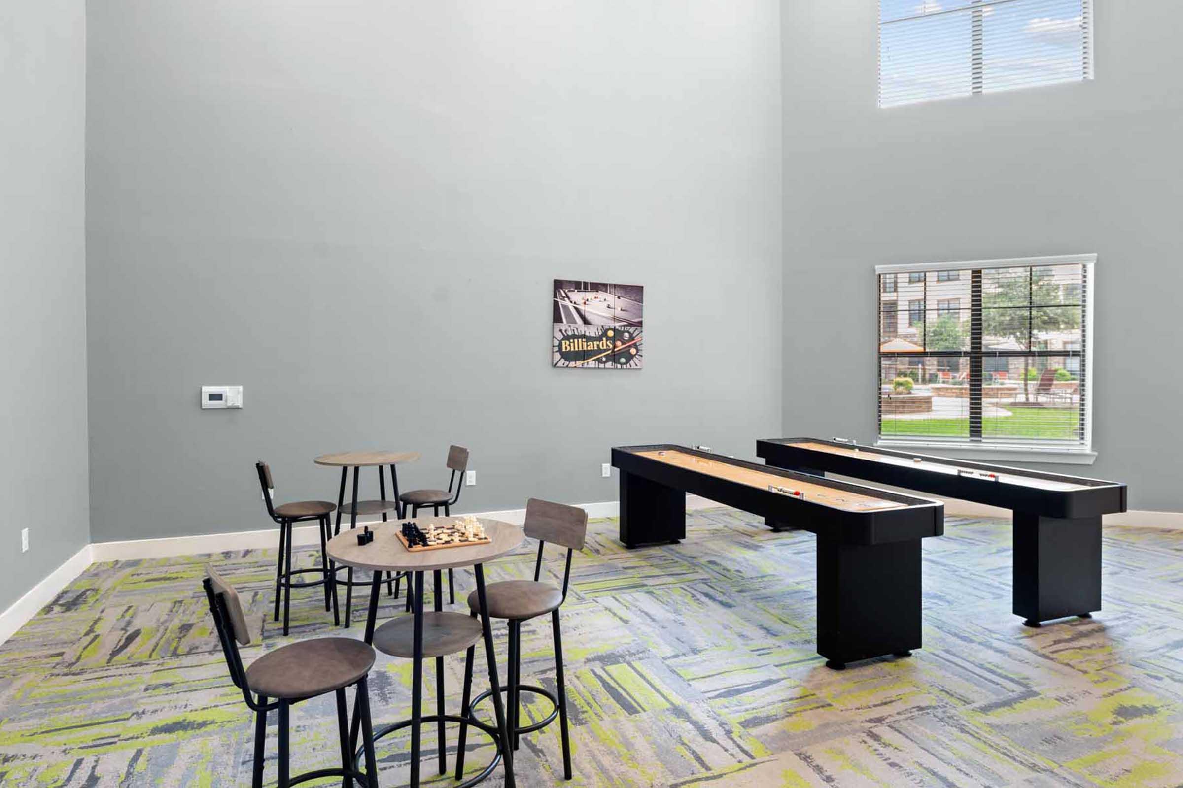 A spacious recreational area featuring a shuffleboard table, a small round table with four chairs, and a wall art piece related to billiards. Large windows allow natural light to fill the room, and the floor is covered with a colorful patterned carpet.