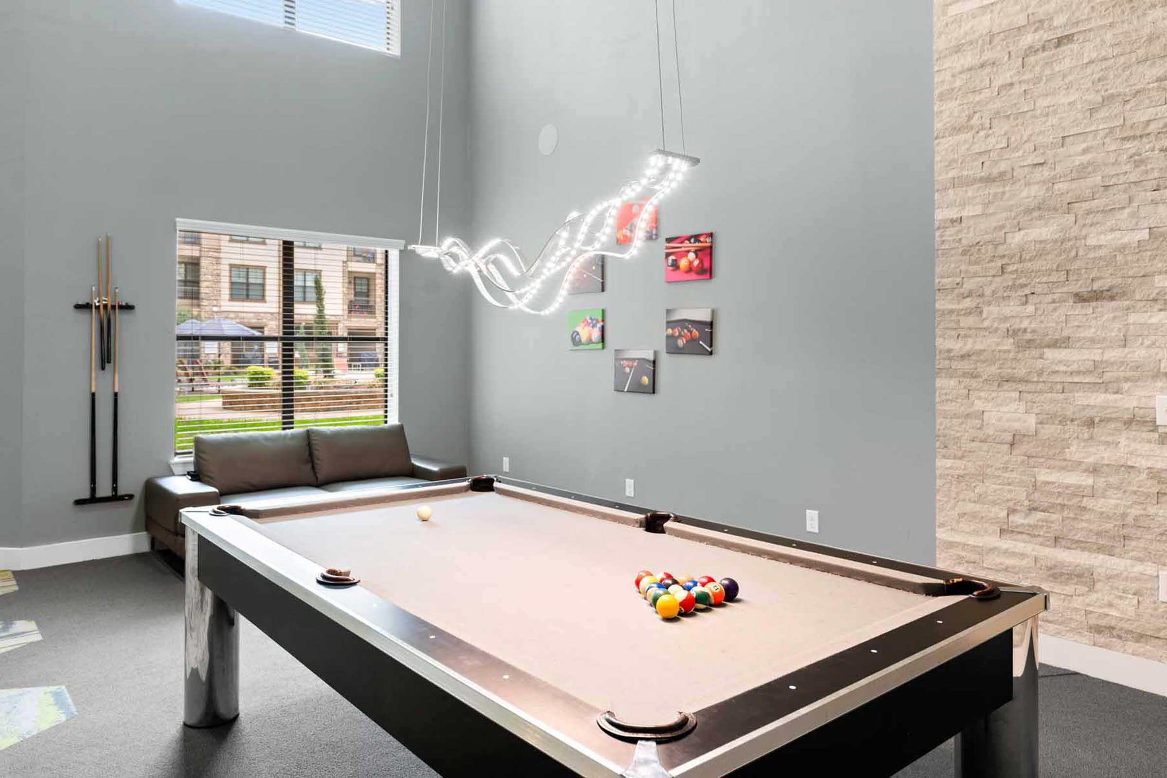 A modern billiard room featuring a sleek pool table with balls arranged for play. Natural light streams through a window, illuminating a cozy seating area with a couch. Stylish lighting fixtures hang above the table, and framed photos decorate the walls, adding an artistic touch to the space.
