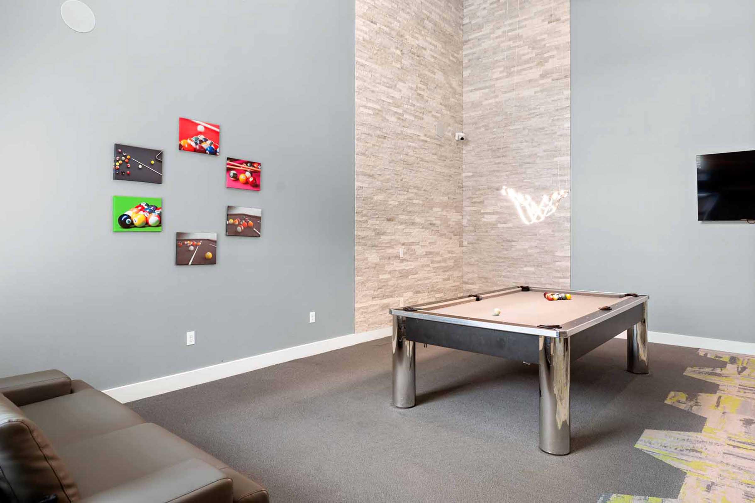A modern game room featuring a sleek pool table with metallic legs, a gray carpet, and a comfortable black couch. On the wall, there are colorful art pieces depicting billiard games. A flat-screen TV is mounted on the wall, and natural light illuminates the space.
