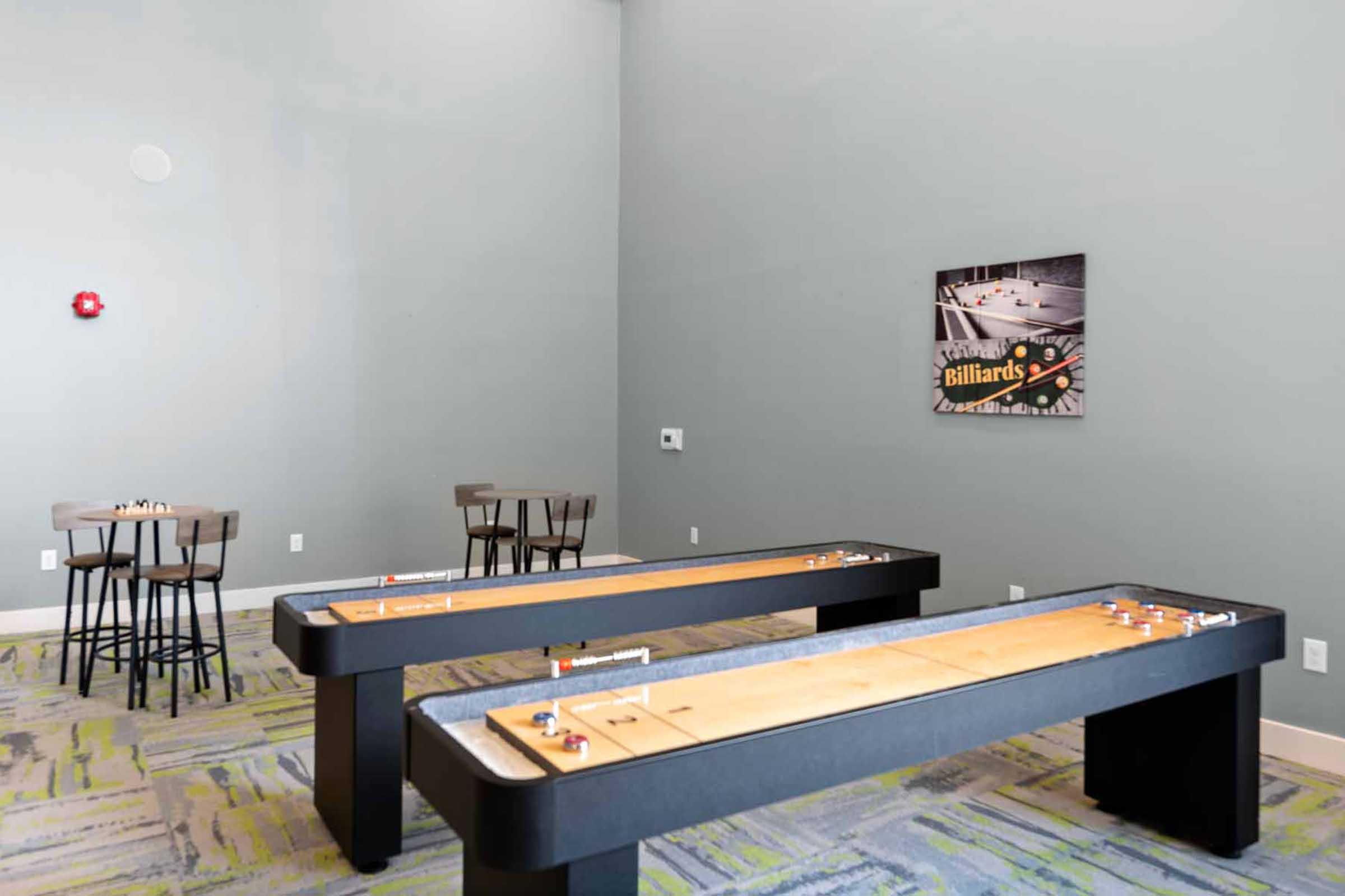 Two shuffleboard tables are positioned in a room with gray walls and a colorful, patterned carpet. There are bar-height tables and chairs nearby. A framed picture of a billiards scene hangs on the wall, adding to the leisure atmosphere.