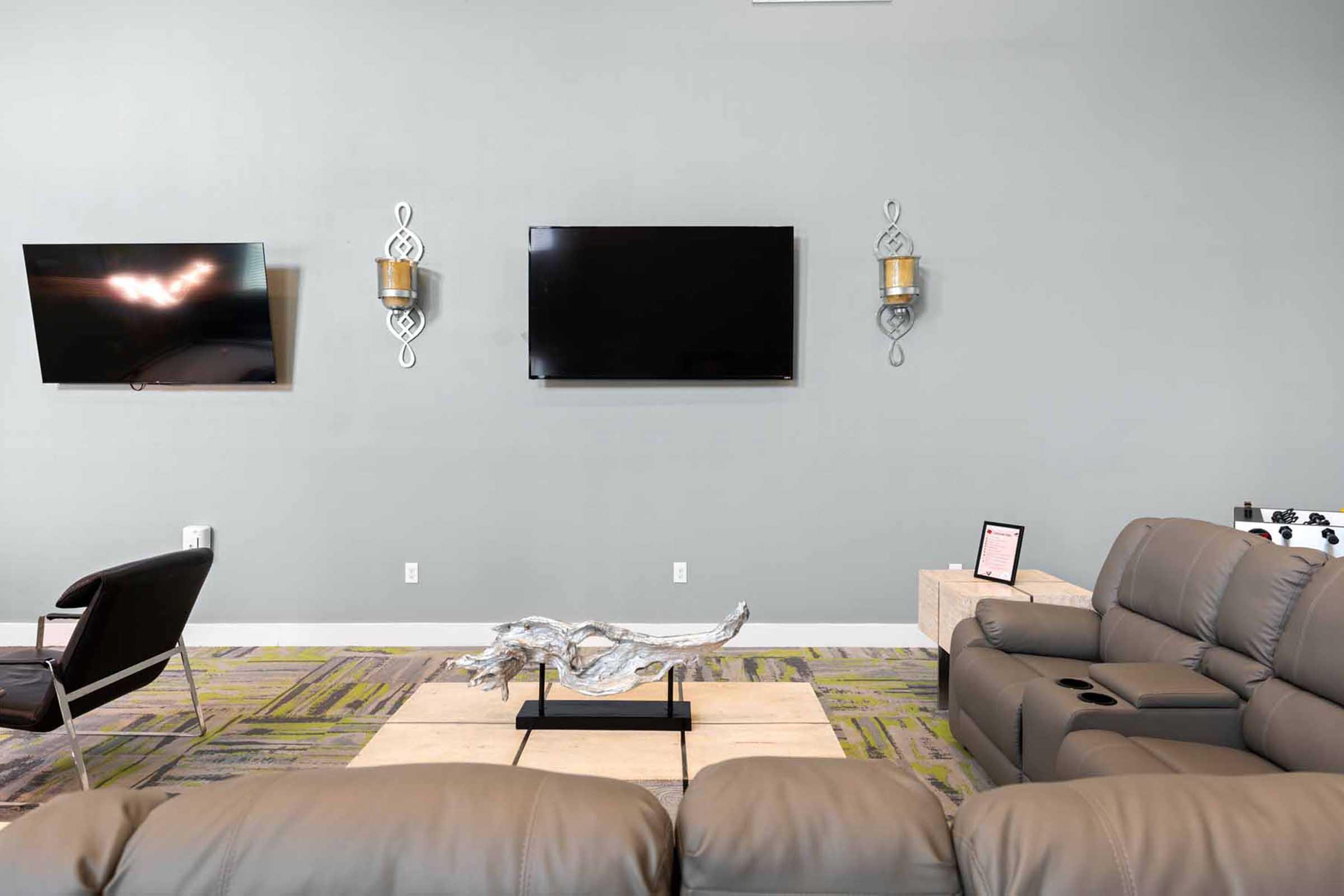 A modern living room with two wall-mounted flat-screen TVs. The seating includes a large gray sofa and a black chair. There’s a coffee table with an artistic wooden centerpiece, and the walls are painted light gray. The flooring features a patterned carpet in shades of green and beige.