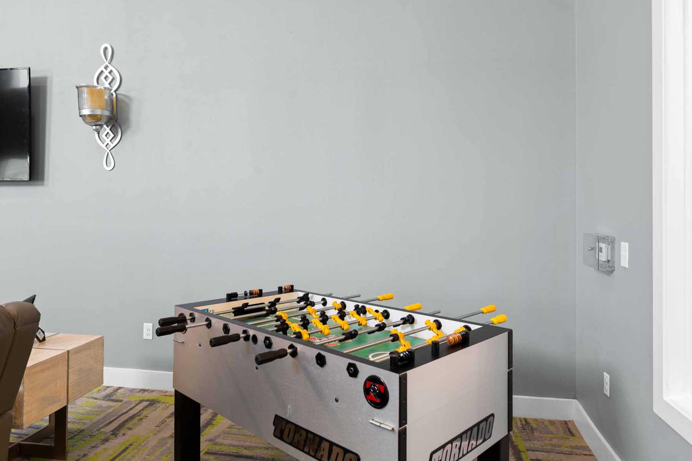 A foosball table in a modern room with gray walls. The table features yellow and black players and is positioned near a wall-mounted light fixture. The room has a cozy atmosphere, with a patterned carpet and minimalistic decor, creating a suitable space for leisure and socializing.