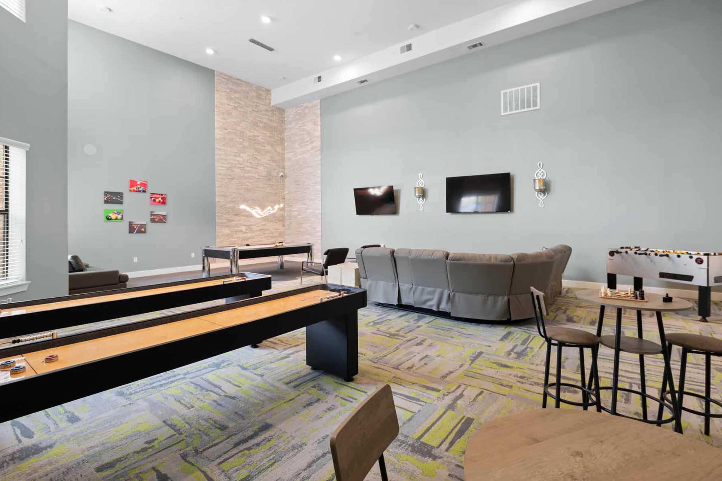 A modern game room featuring a shuffleboard table, a comfortable seating area with a gray sectional couch, and a wall displaying various framed artworks. Two wall-mounted TVs are visible, along with small tables and chairs, creating an inviting space for leisure and entertainment.