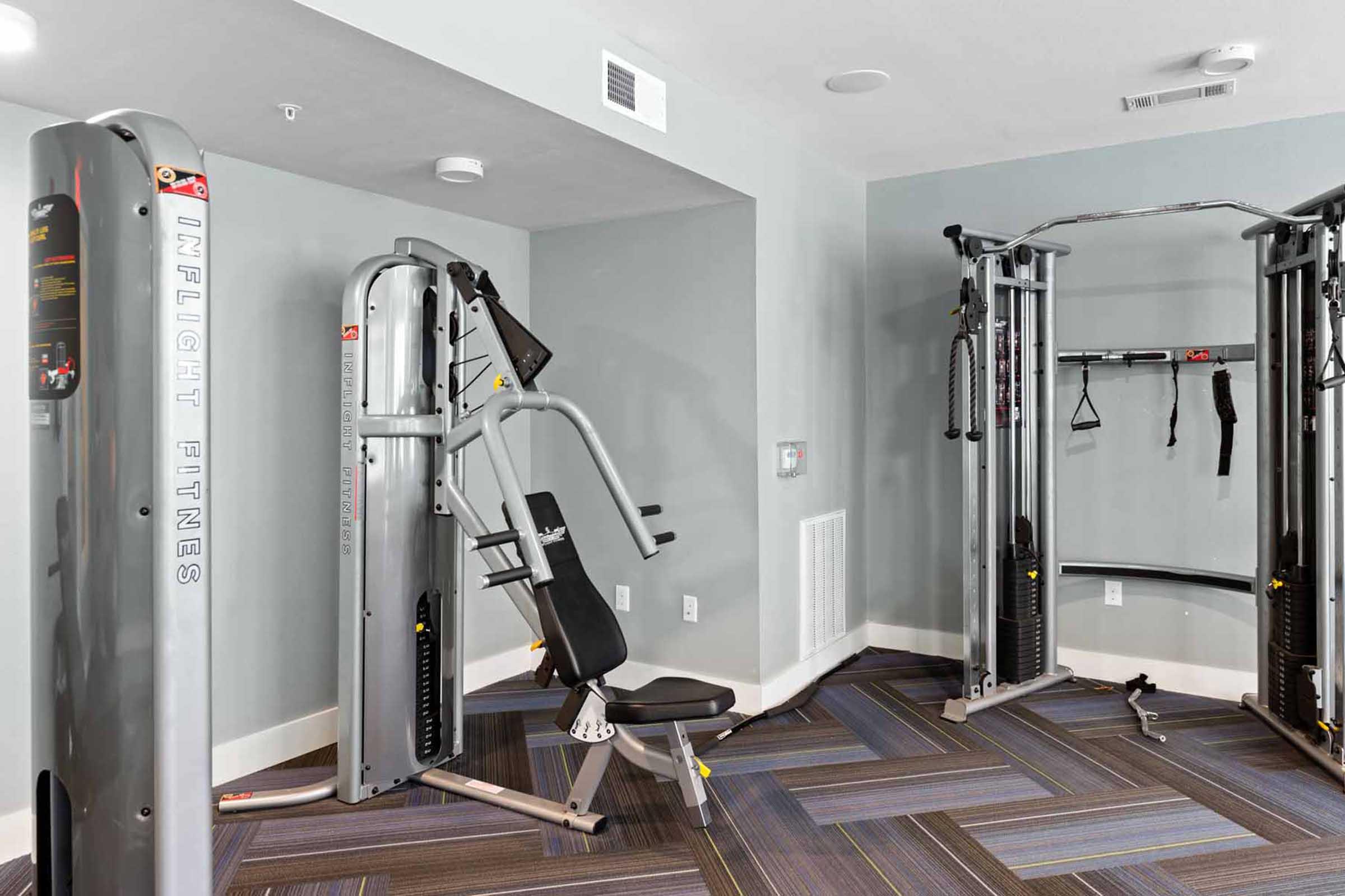 A modern fitness room featuring two multifunctional gym machines for strength training. One machine is designed for upper body workouts, while the other includes various attachments for versatility. The space has light-colored walls and a patterned carpet, creating a clean and inviting atmosphere.