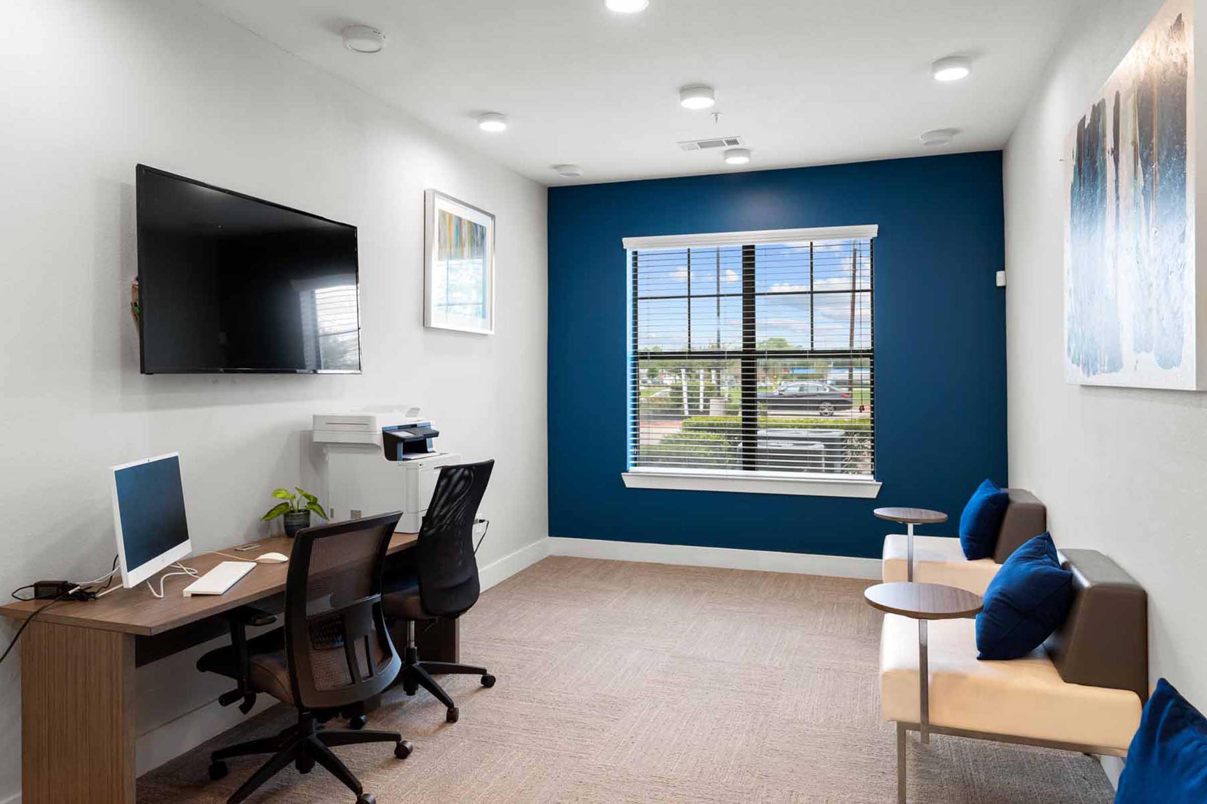 A modern office space featuring a desk with a computer, a television mounted on the wall, and two comfortable chairs with small side tables. The room has a large window with blinds, allowing natural light, and is decorated with a piece of artwork on the wall. The color scheme includes blue and neutral tones.