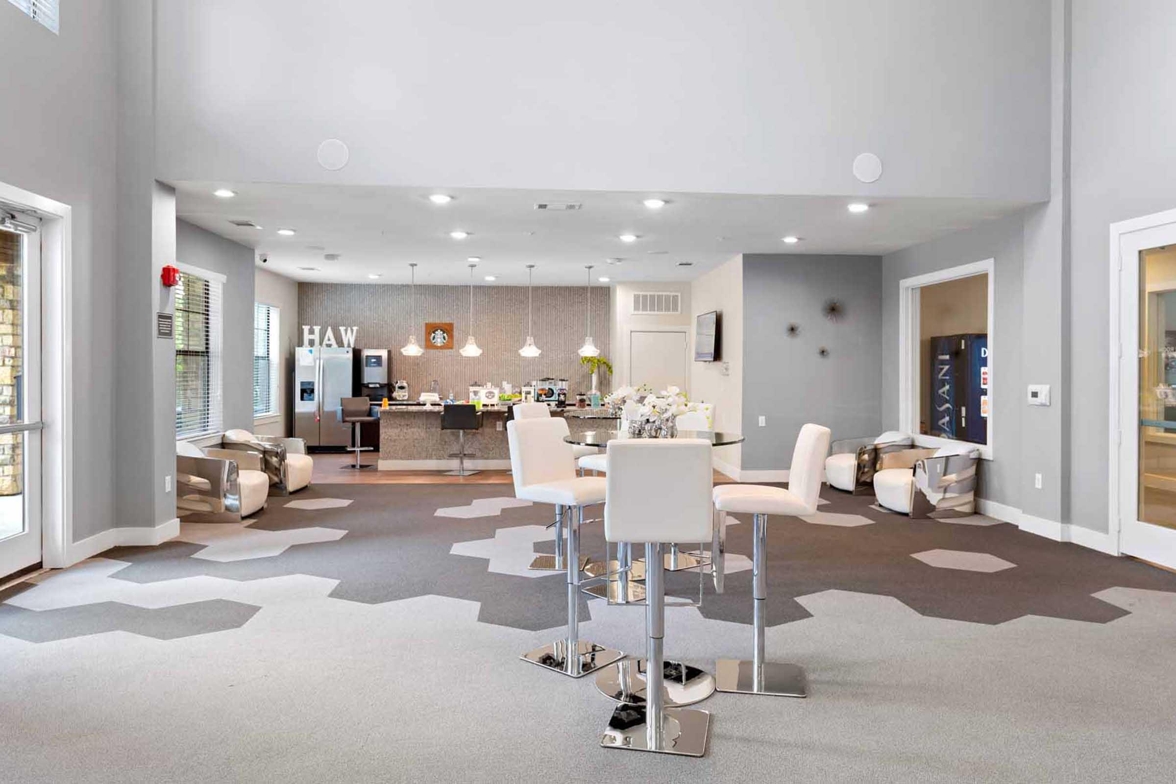 Modern interior of a spacious lounge area featuring light gray walls, stylish furniture, and decorative hexagonal carpet. A coffee bar is visible in the background with seating and decor. Large windows allow natural light to flood the space, creating an inviting atmosphere for social gatherings.