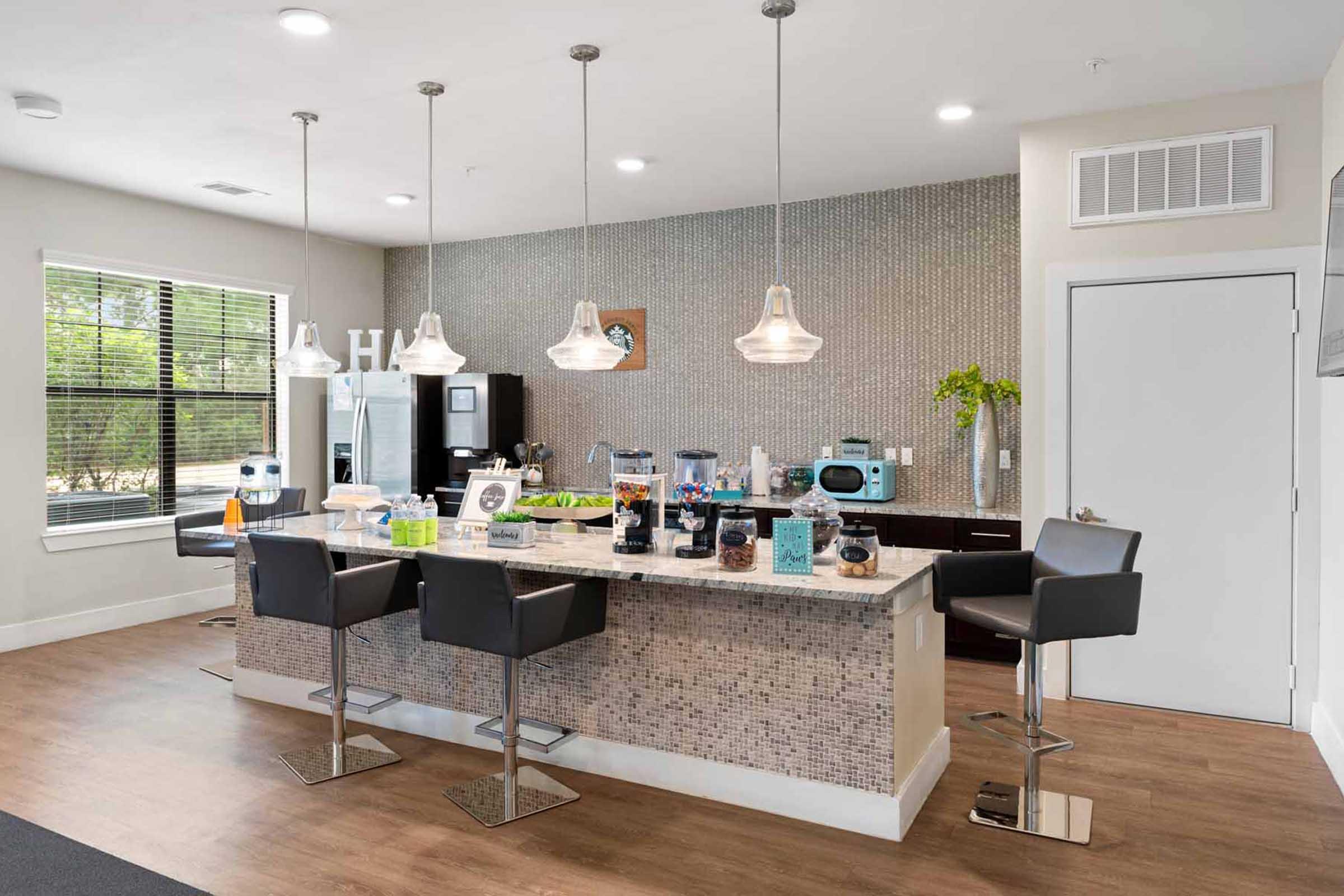 A modern kitchen or bar area featuring a sleek countertop with bar stools, pendant lighting, and a mosaic backsplash. The space includes various kitchen appliances, decorative items, and a green plant, creating a contemporary and inviting atmosphere.
