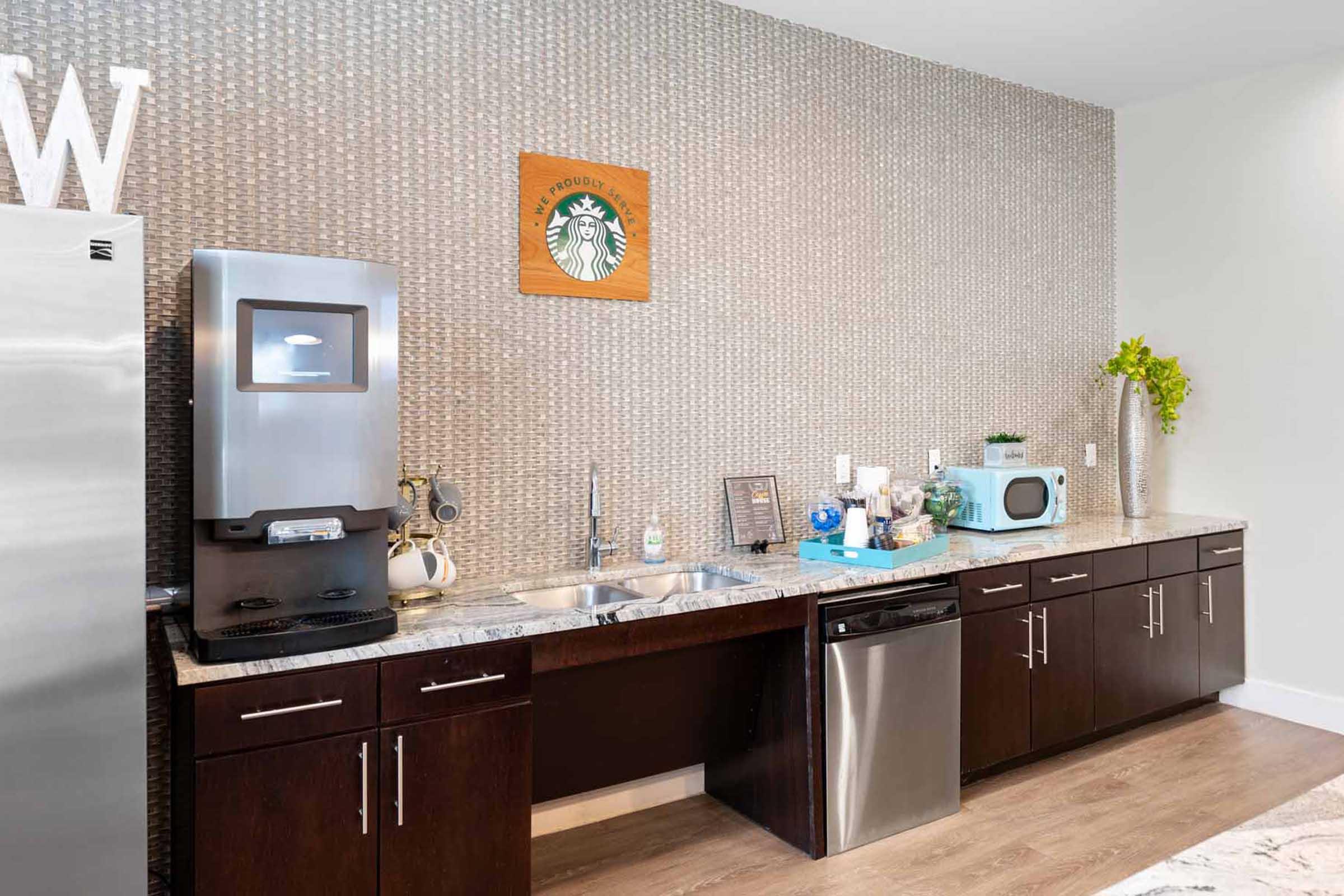 Modern kitchen area featuring a textured wall, a stainless steel refrigerator, a countertop with a double sink, and a Starbucks-themed wall art. There's a coffee machine, a microwave, and decorative items on the counter, with dark wood cabinetry and light wooden flooring.