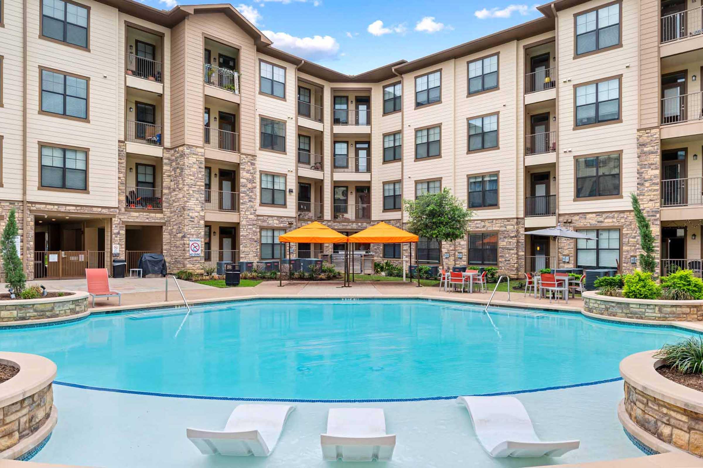 A well-maintained apartment complex featuring a swimming pool surrounded by lounge chairs, umbrellas, and patio seating. The buildings are designed with a mix of stone and siding, set against a clear blue sky with scattered clouds, creating a relaxing and inviting atmosphere.