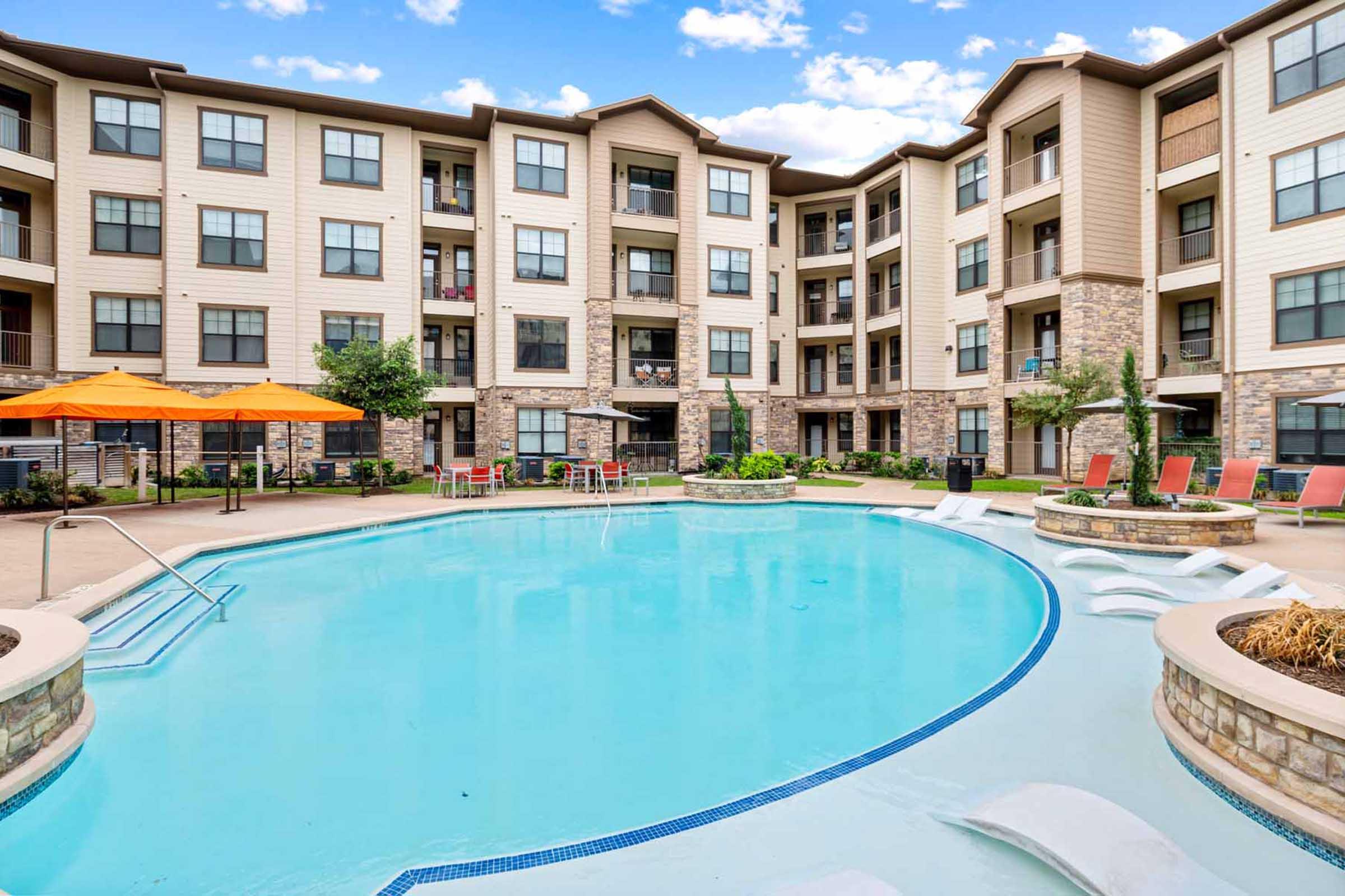 A clear blue swimming pool surrounded by modern apartment buildings. The pool area features lounge chairs and vibrant orange umbrellas, with landscaped greenery adding to the aesthetic. The sky above is partly cloudy, enhancing the inviting atmosphere of the outdoor space.