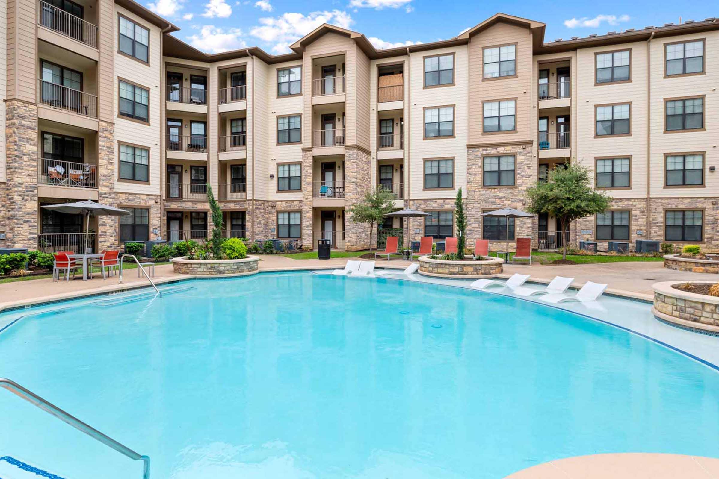 An outdoor swimming pool surrounded by a landscaped courtyard of a multi-story apartment complex. The pool features lounge chairs and is flanked by trees and shrubs, with balconies visible on the building exterior. Clear blue skies and fluffy clouds are overhead.