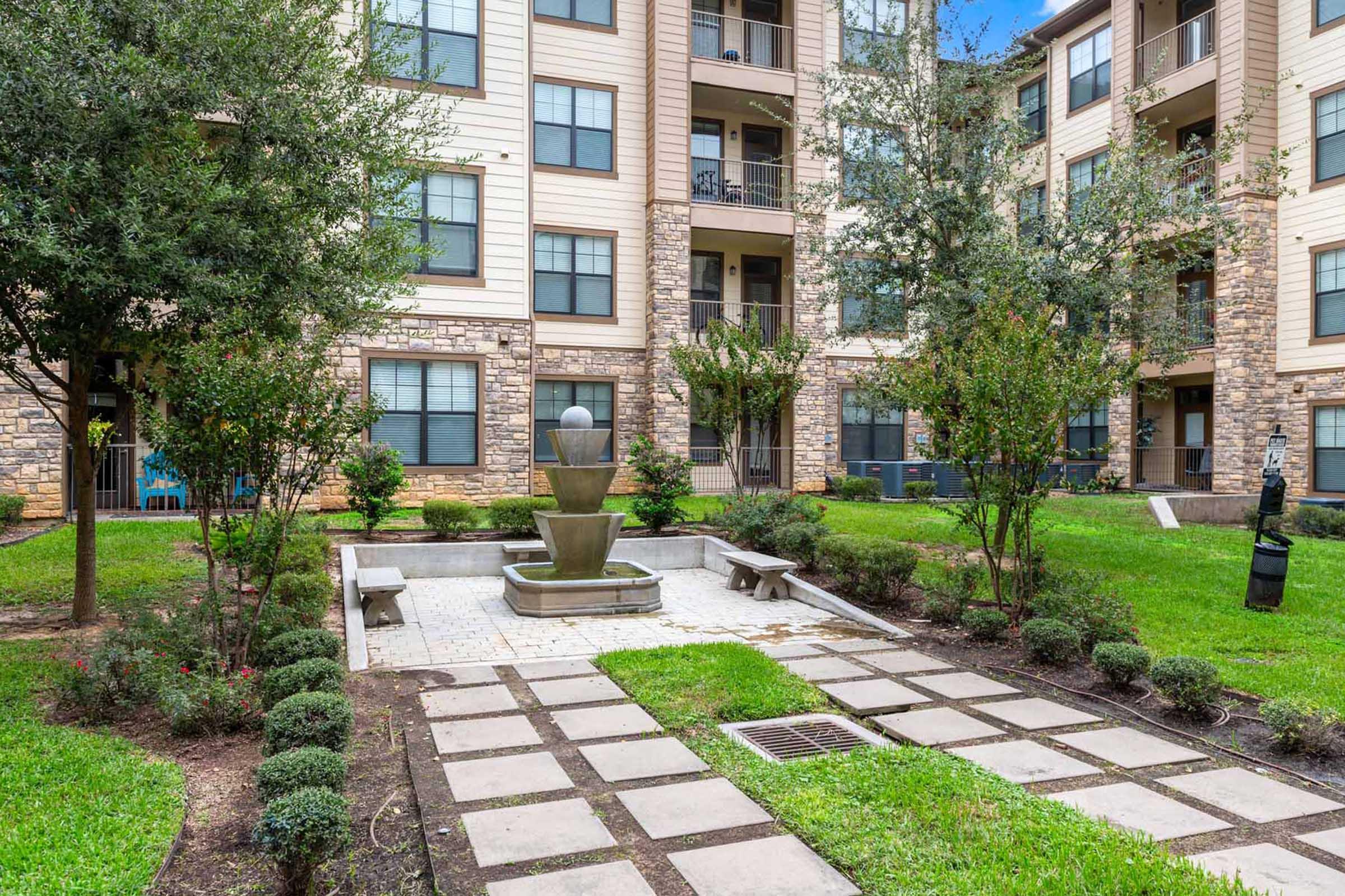 A peaceful courtyard featuring a tiered fountain surrounded by neatly trimmed bushes, benches, and stone pathways. Several apartment buildings with balconies and windows are visible in the background, offering a serene atmosphere for residents and visitors.