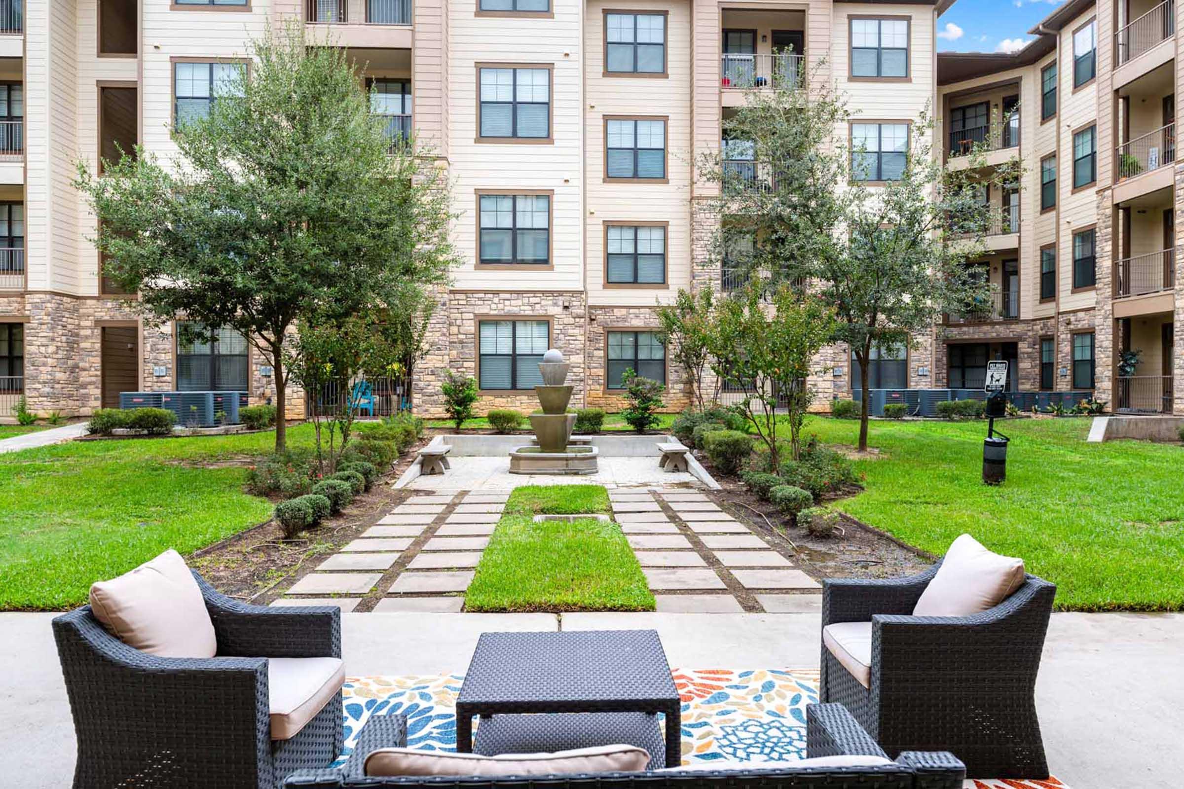 A peaceful outdoor space featuring a small seating area with two chairs and a table, surrounded by lush green grass and neatly trimmed bushes. In the background, there are several apartment buildings, and a decorative fountain is centered in a garden pathway.